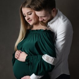 Kamila Jankowska Fotografia - Ciąża: Kobieta w szmaragdowej sukni z koronkowymi rękawami, obejmowana przez mężczyznę na ciemnym tle, oboje spoglądają na brzuch.