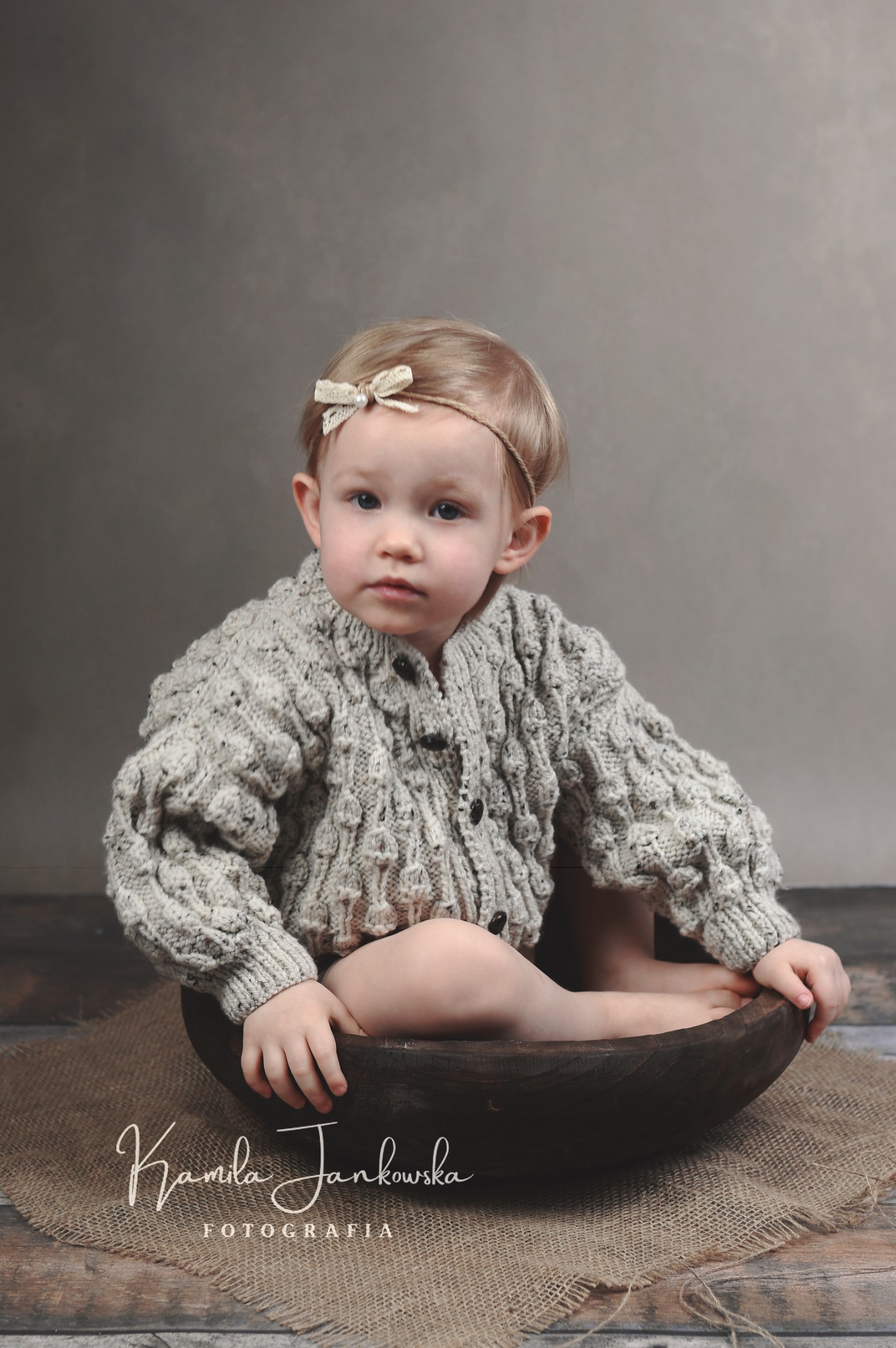 Mała dziewczynka w grubym swetrze i opasce z kokardką siedzi w drewnianej misce na jutowym materiale, portret w neutralnej tonacji.
