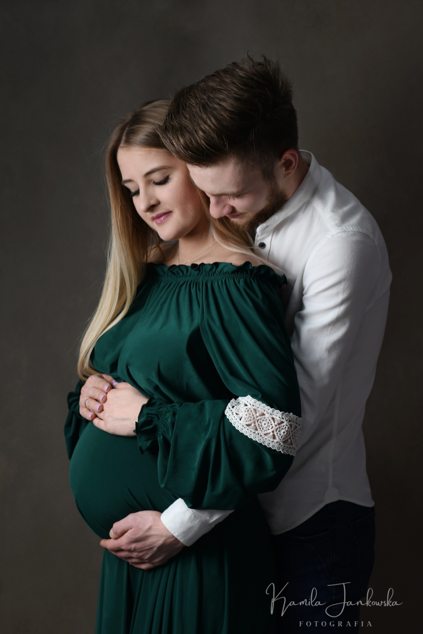 Ciąża: Kobieta w szmaragdowej sukni z koronkowymi rękawami, obejmowana przez mężczyznę na ciemnym tle, oboje spoglądają na brzuch.