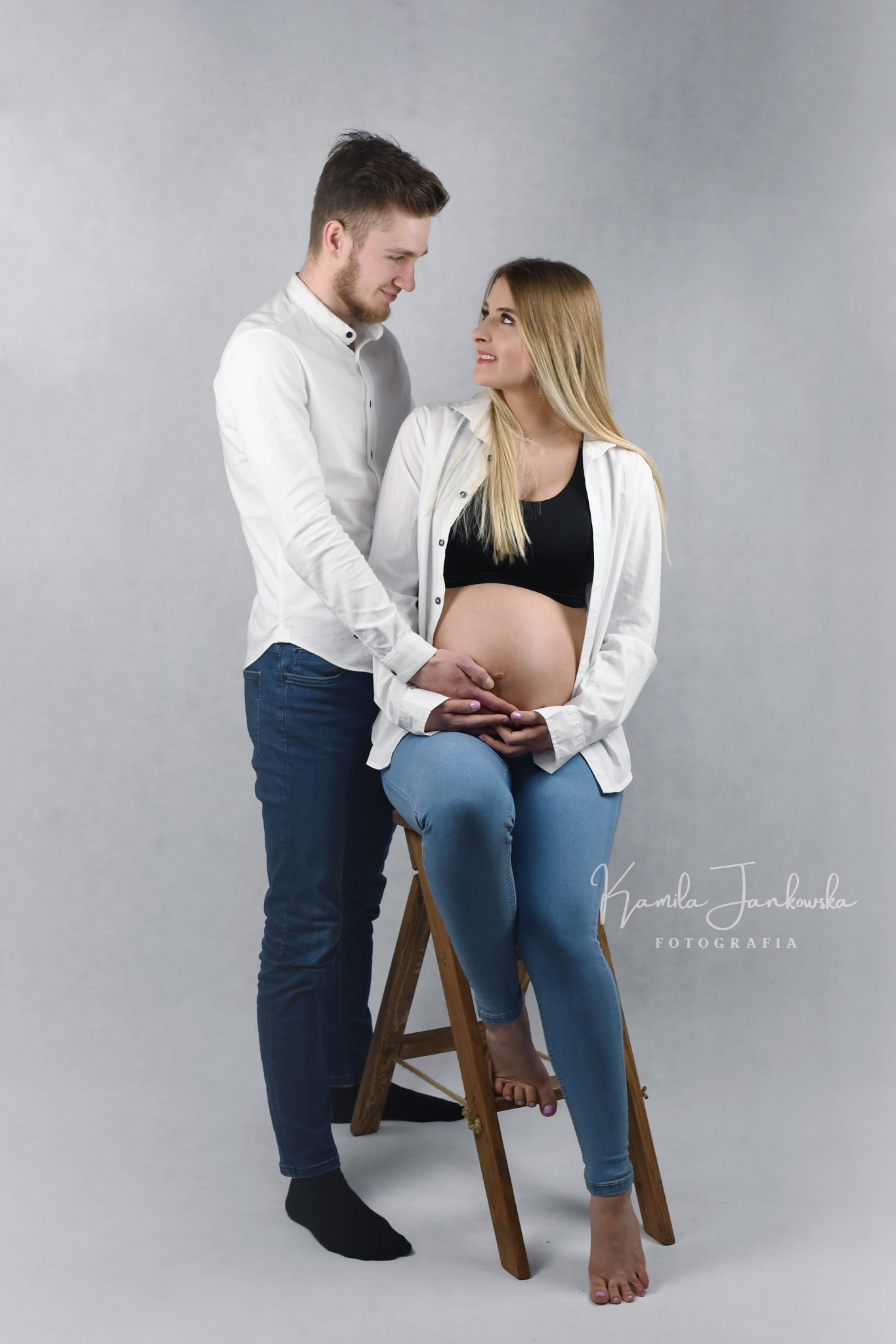 Ciężarna kobieta siedzi na drewnianym stołku, ubrana w jeansy i rozpiętą białą koszulę, partner stoi za nią, oboje patrzą na siebie z uśmiechem, dłonie mężczyzny spoczywają na brzuchu kobiety...