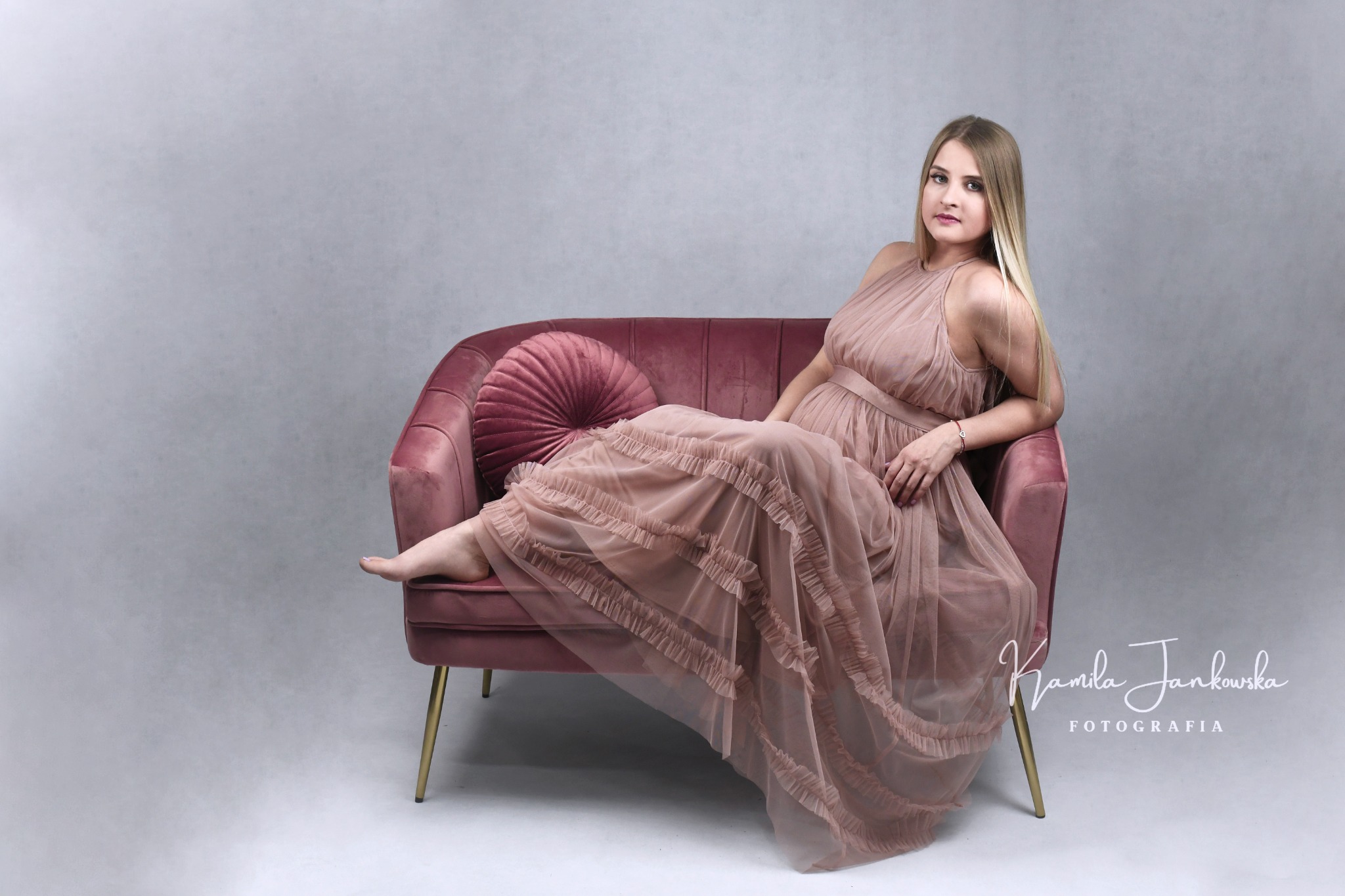 Kobieta w zwiewnej sukni ciążowej pozuje siedząc na różowym welurowym fotelu z okrągłą poduszką na tle szarego studia fotograficznego.