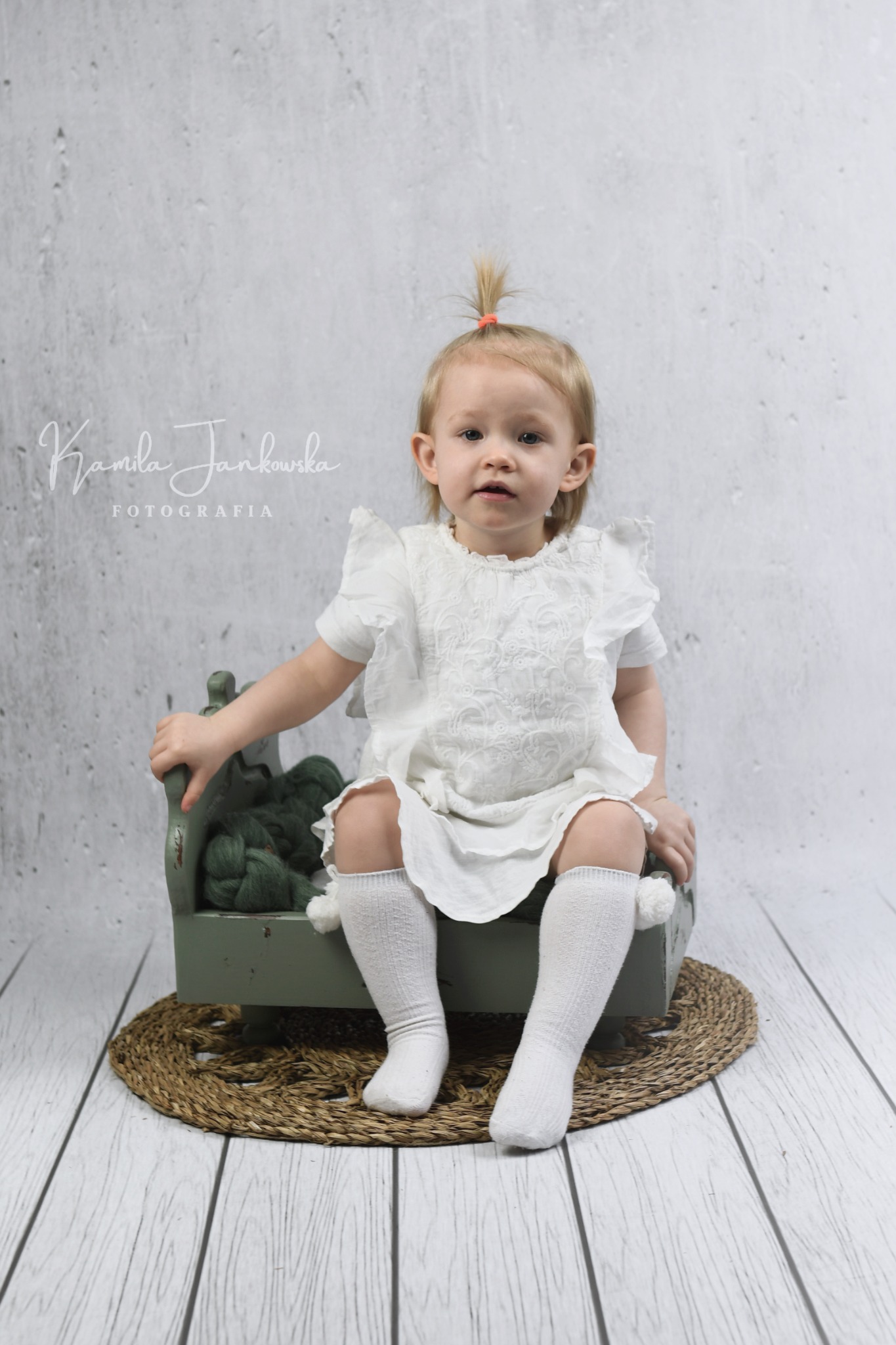 Dziewczynka w białej sukience siedzi na małym, zielonym łóżeczku z wełnianym wypełnieniem na tle drewnianej podłogi i szarej ściany z widocznym logo fotografa.
