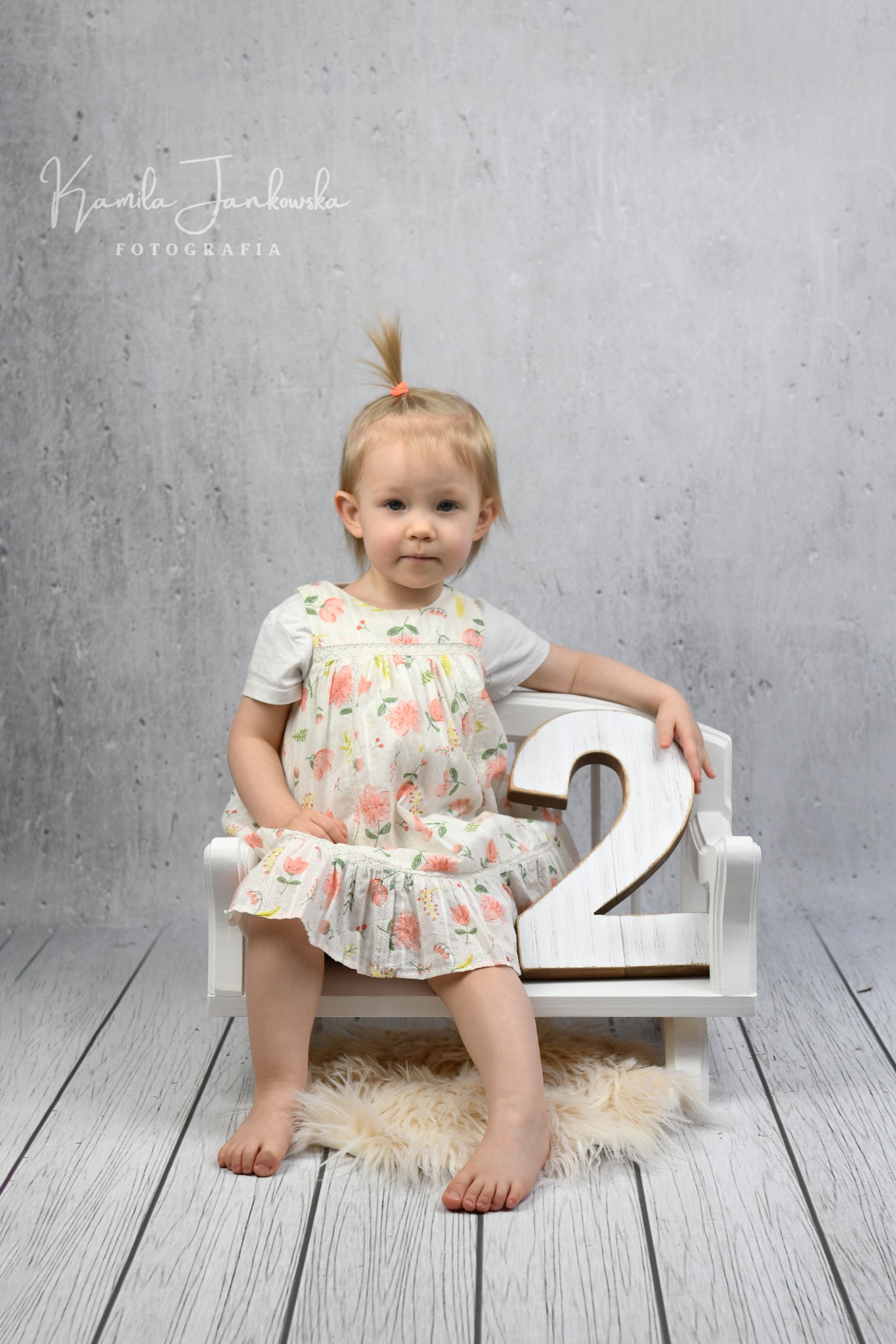 Dziewczynka z blond włosami siedzi na małym białym krześle, trzymając drewnianą cyfrę 2, na tle szarej ściany i drewnianej podłogi.