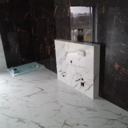 Łazienka w trakcie wykańczania: ułożone białe płytki podłogowe z marmurkowym wzorem, ciemna ściana z płytek imitujących marmur, oraz biała zabudowa podtynkowego systemu WC z przygotowanymi otworami...