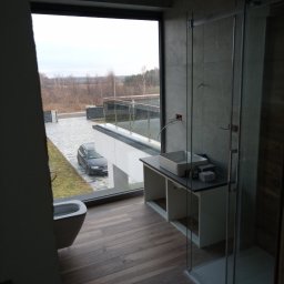 Janusz Sposób - Nowoczesna łazienka z dużym oknem wychodzącym na podjazd, widoczna szara umywalka nablatowa, kabina prysznicowa ze szklanymi drzwiami i drewnopodobna podłoga.