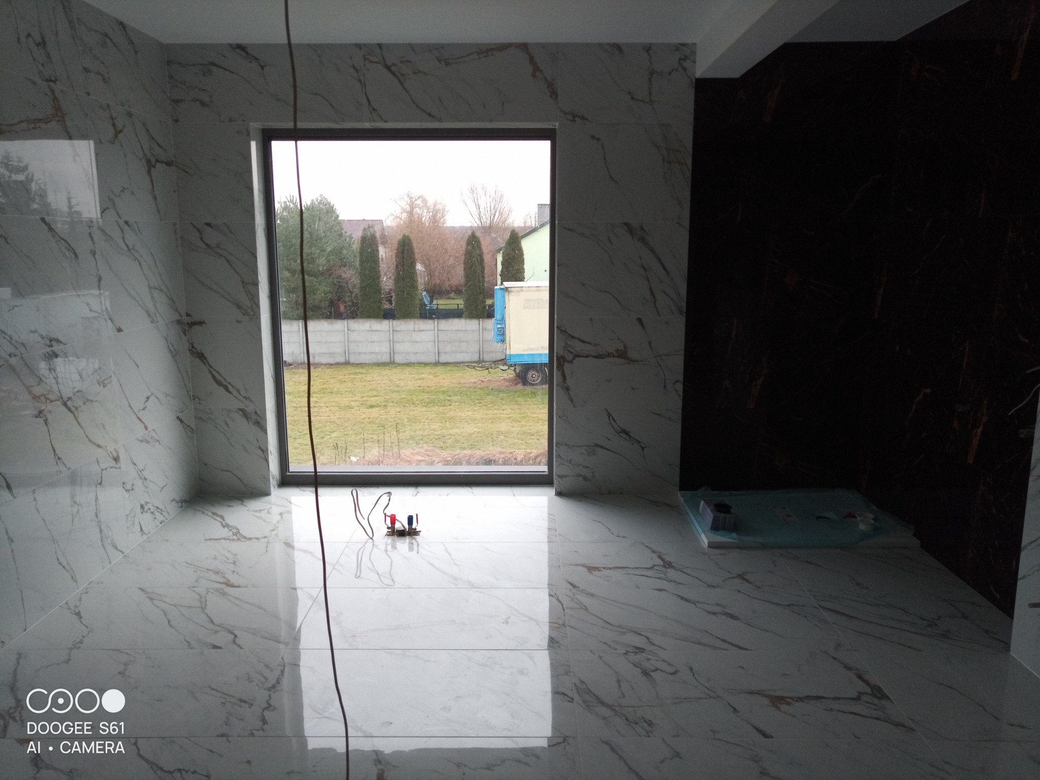 Wnętrze łazienki w trakcie wykańczania z białymi płytkami imitującymi marmur na ścianach i podłodze, widoczne okno z widokiem na ogród oraz czarna ściana z ciemnymi płytkami, wystające kable...