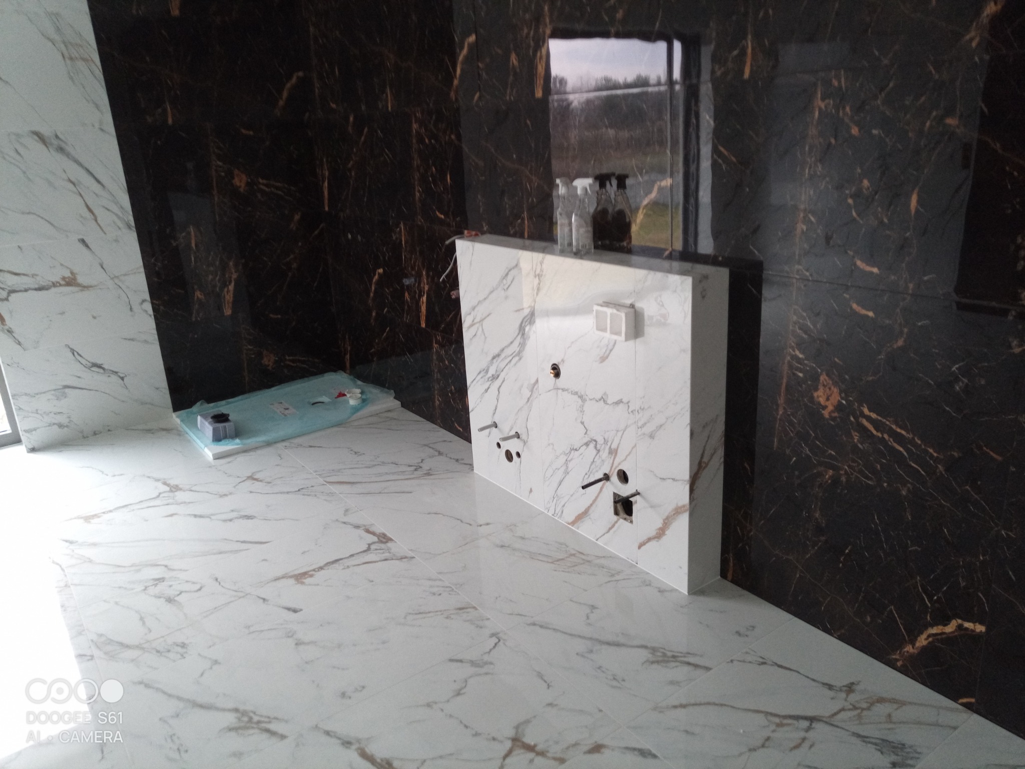 Łazienka w trakcie wykańczania: ułożone białe płytki podłogowe z marmurkowym wzorem, ciemna ściana z płytek imitujących marmur, oraz biała zabudowa podtynkowego systemu WC z przygotowanymi otworami...