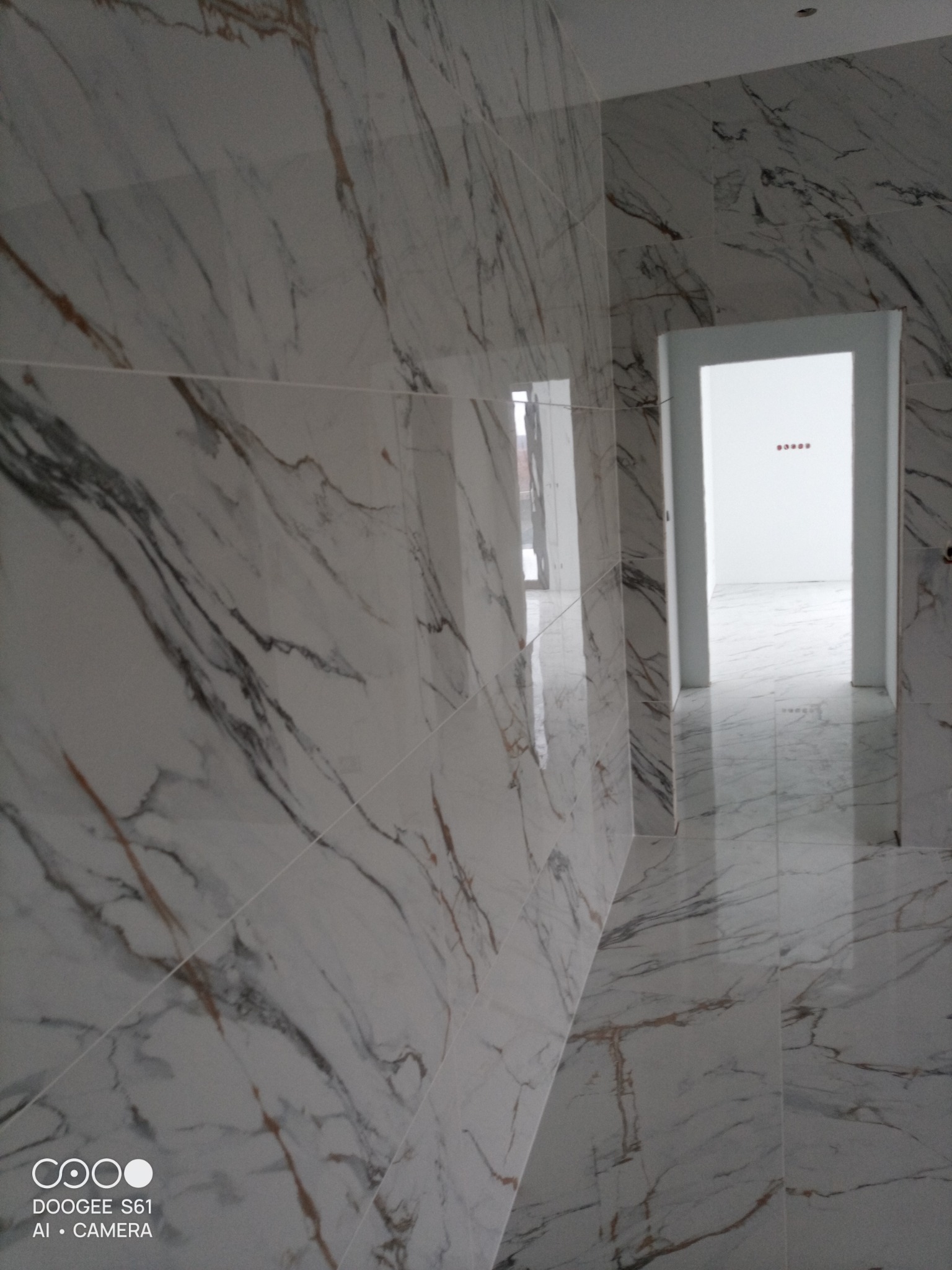 Wykończone wnętrze łazienki z białymi płytkami imitującymi marmur na ścianach i podłodze, widok z perspektywy, otwarte drzwi do kolejnego pomieszczenia z widocznymi puszkami elektrycznymi.