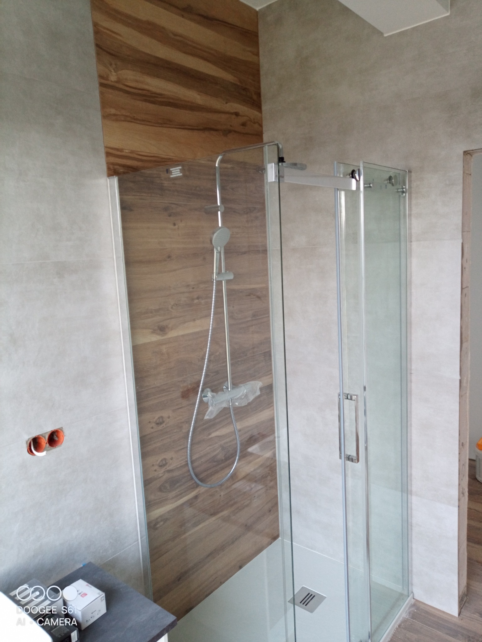 Nowoczesna kabina prysznicowa z drewnopodobną ścianą, widoczny odpływ liniowy i bateria prysznicowa z deszczownicą, szare płytki na ścianach.