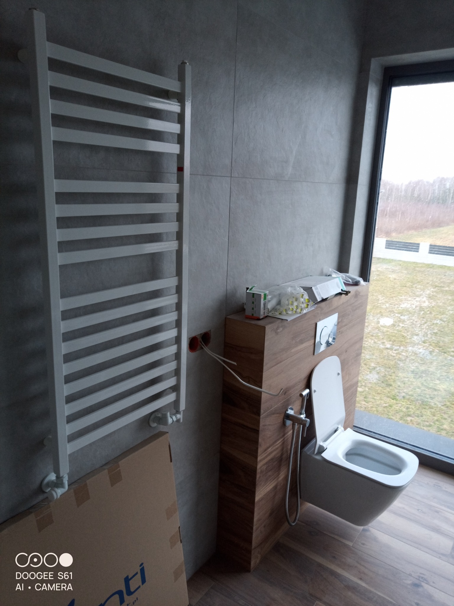 Nowoczesna łazienka z podwieszaną toaletą, bidetką i grzejnikiem drabinkowym, wykończona szarymi płytkami i drewnopodobną zabudową, widok na zewnątrz przez duże okno.