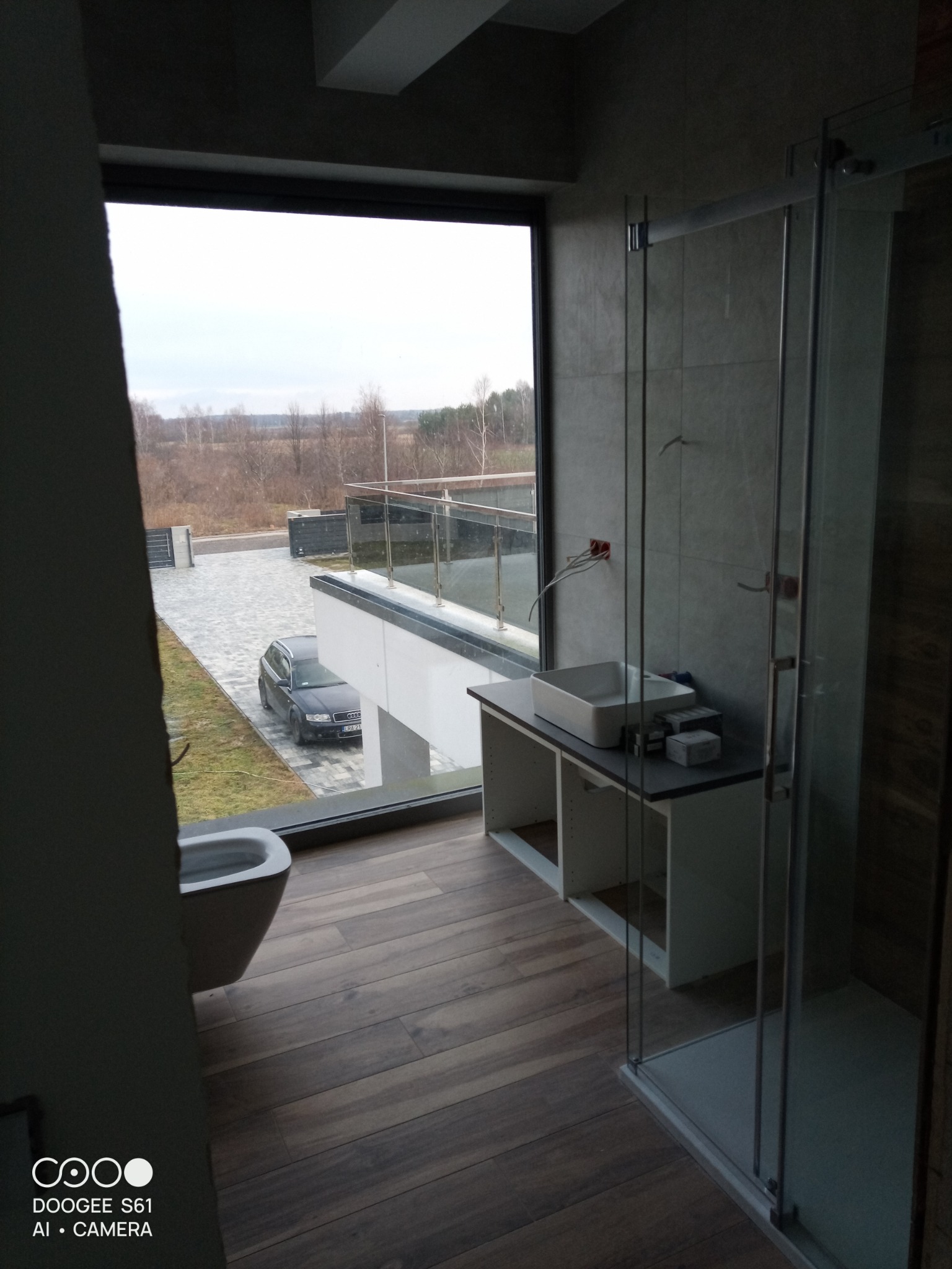 Nowoczesna łazienka z dużym oknem wychodzącym na podjazd, widoczna szara umywalka nablatowa, kabina prysznicowa ze szklanymi drzwiami i drewnopodobna podłoga.