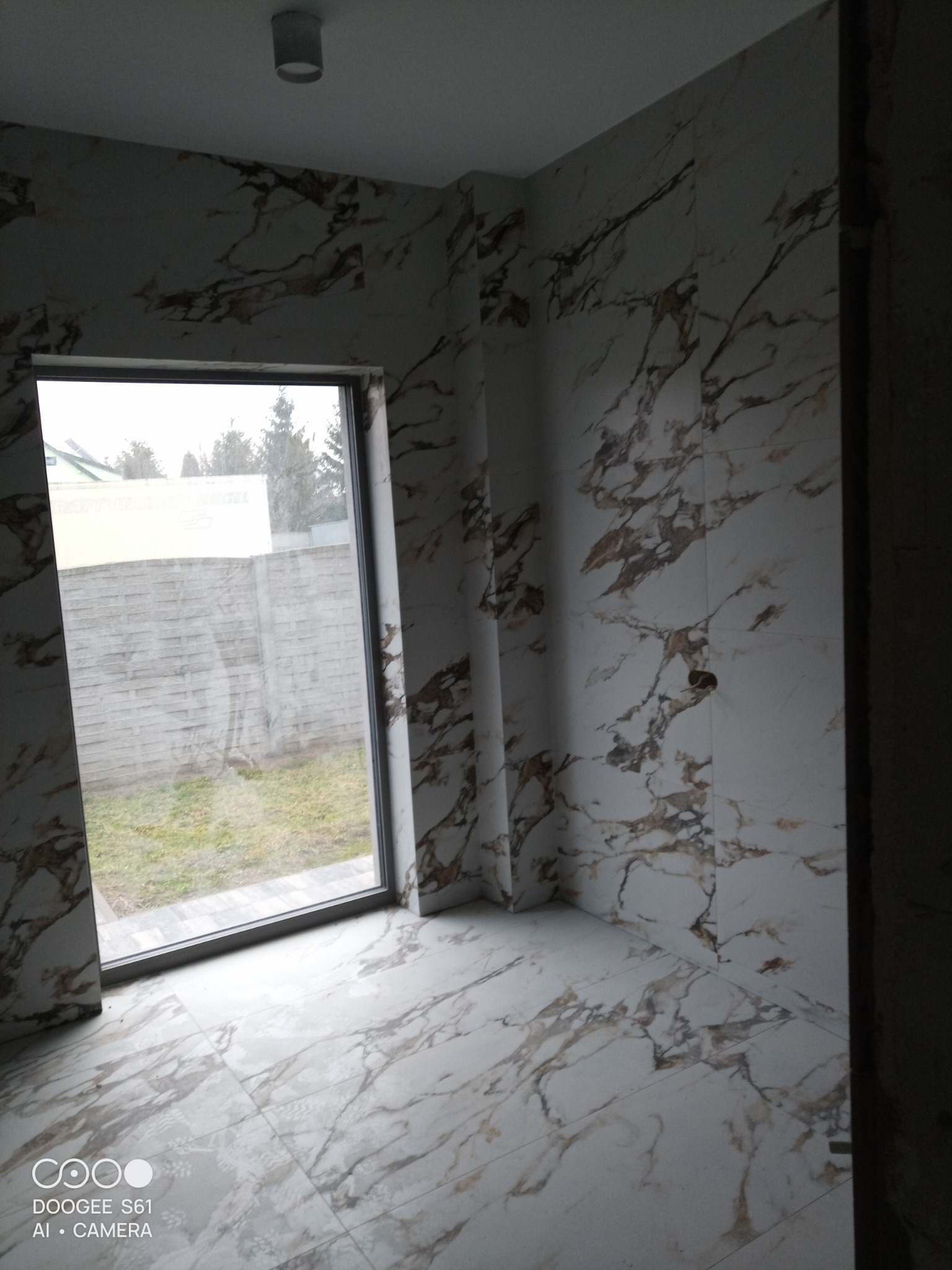 Wykończona łazienka z płytkami imitującymi marmur na ścianach i podłodze, widok na szary betonowy płot przez okno.