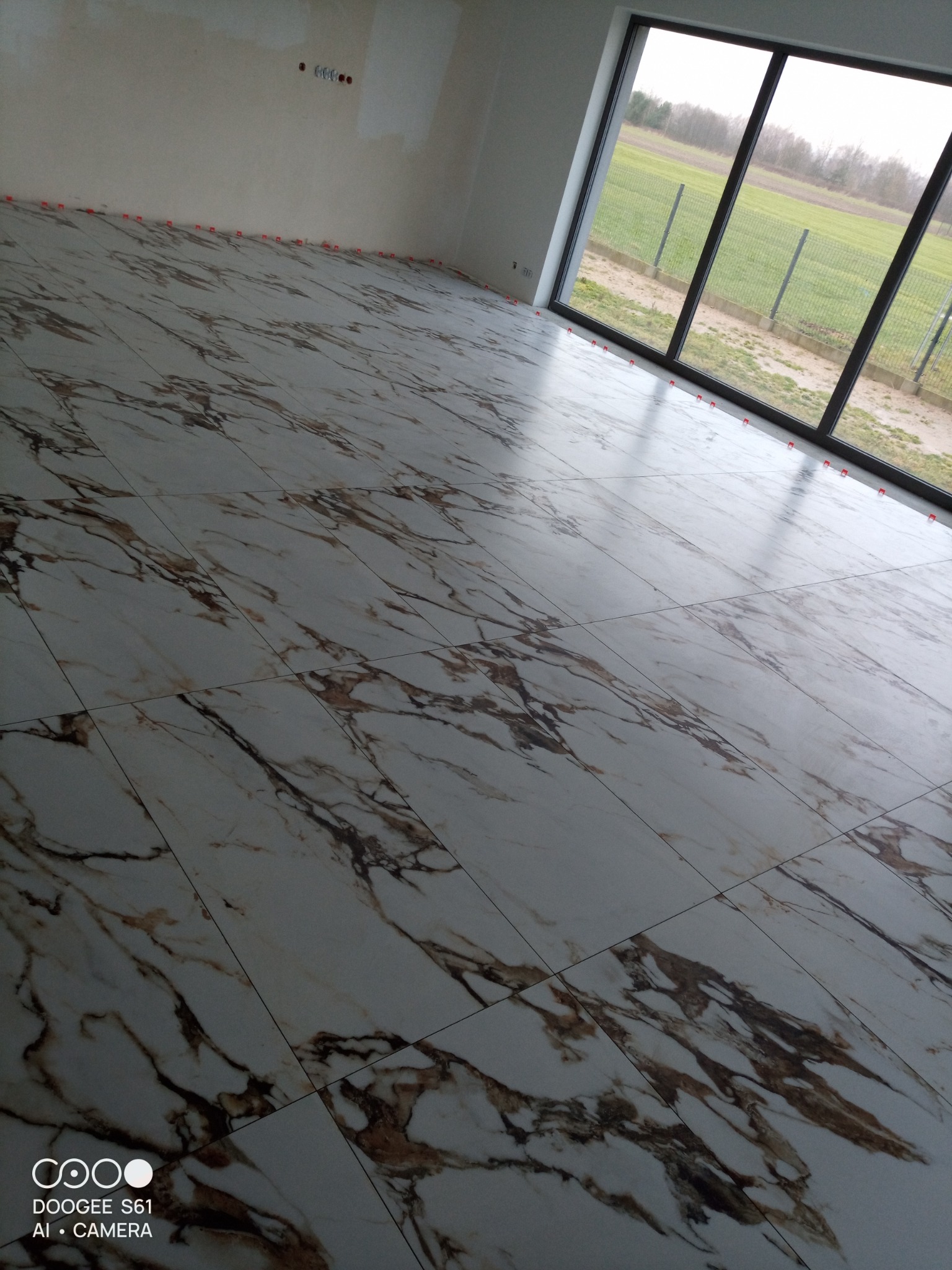 Ułożone płytki podłogowe imitujące marmur w jasnym pomieszczeniu z dużym oknem wychodzącym na zielone pole; widoczne plastikowe krzyżyki dystansowe między płytkami.
