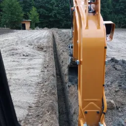 Pomarańczowa koparka z widocznymi elementami hydraulicznymi zagłębia się w ziemię, tworząc wąski wykop w piaszczystym terenie. W tle widoczny mały budynek i drzewa.