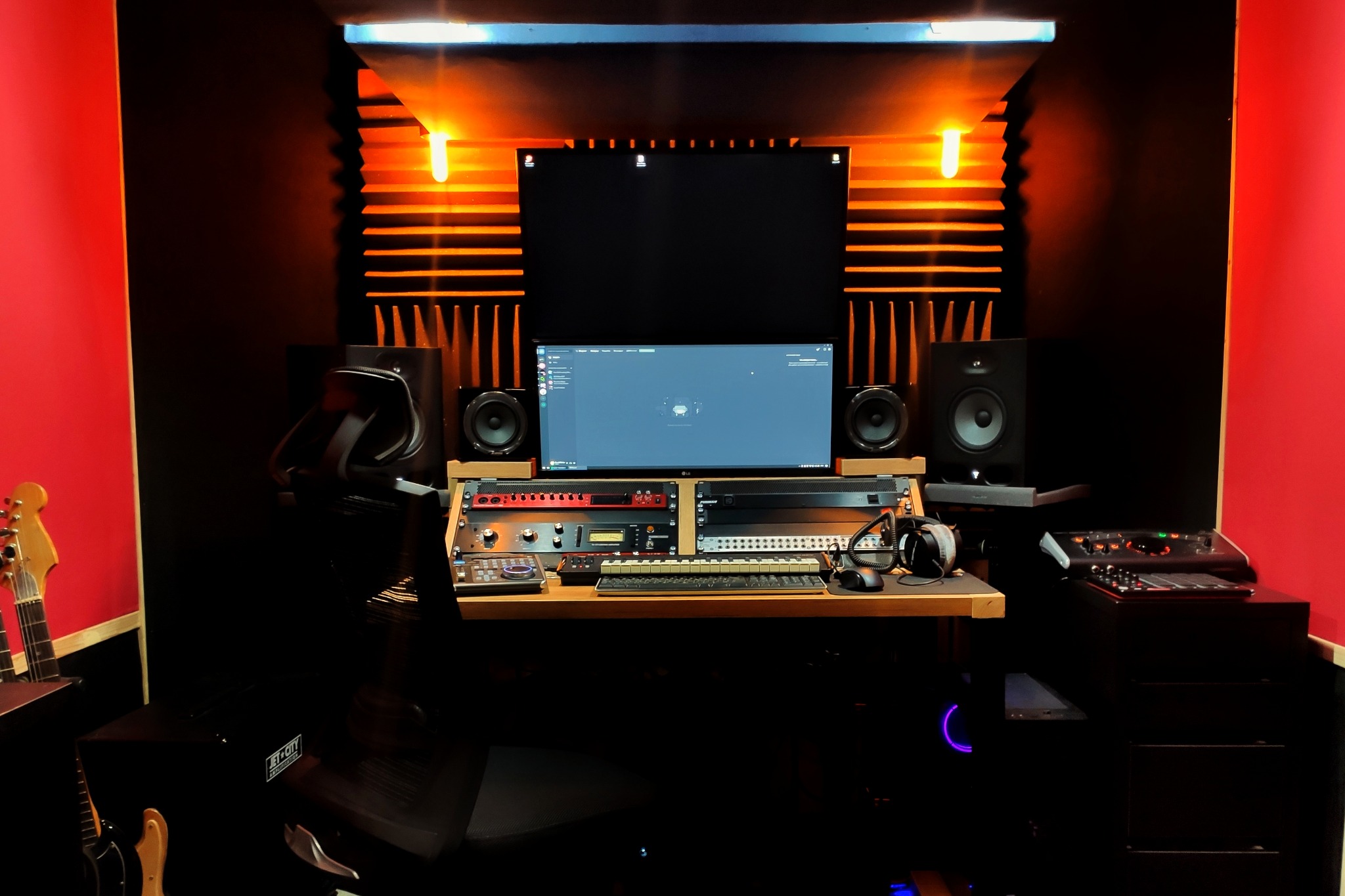 Profesjonalne studio nagrań: monitory studyjne, dwa ekrany, konsoleta mikserska, klawiatura MIDI, słuchawki studyjne, gitary elektryczne w rogu, panele akustyczne z podświetleniem, czarne ściany...