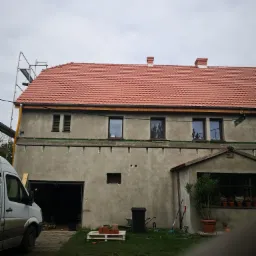 Budynek z szarą elewacją w trakcie prac, widoczne rusztowanie, nowe czerwone dachówki, przygotowane miejsce pod ocieplenie, biały samochód dostawczy z boku