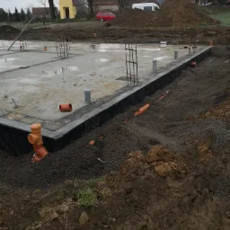 Przygotowany fundament z hydroizolacją pionową i rurami drenażowymi w gruncie, widoczne zbrojenie i betonowa płyta.