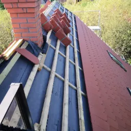 Częściowy widok dachu w trakcie wymiany, z widocznym kominem z czerwonej cegły, łatami, membraną dachową z nadrukiem Wienerberger Plus oraz ułożonymi dachówkami karpiówkami w kolorze ceglastym.