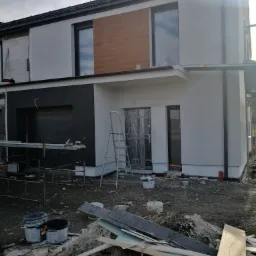 Nowoczesny dom jednorodzinny w trakcie prac wykończeniowych elewacji: fragment otynkowanej ściany, schody przystawne, puste wiadra po farbie i resztki materiałów budowlanych na ziemi.