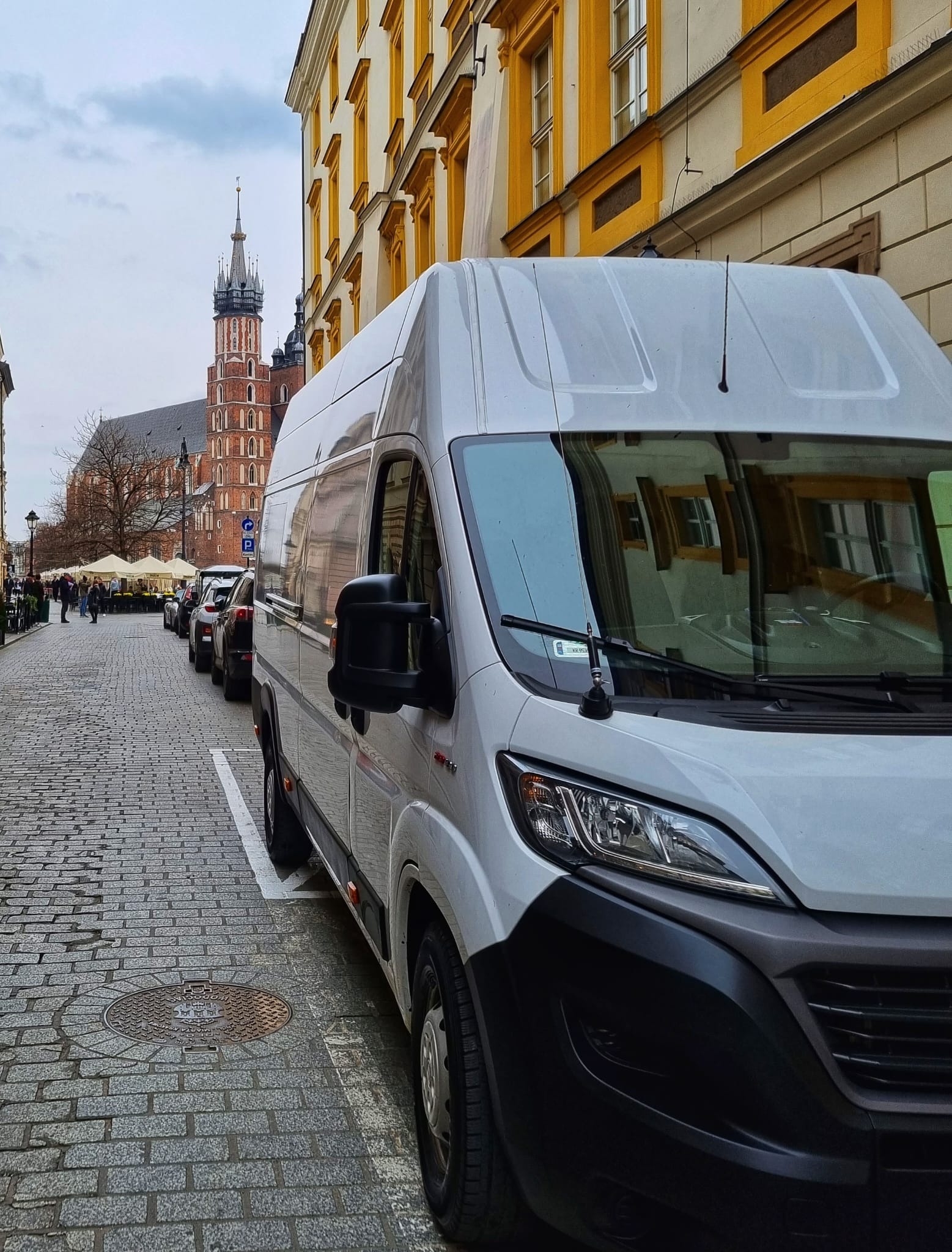 Biały bus zaparkowany na brukowanej ulicy w Krakowie, z widokiem na Bazylikę Mariacką w tle.