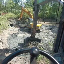 Widok z kabiny maszyny na żółtą koparkę JCB pracującą w wykopie wypełnionym błotnistą ziemią, z widocznymi drzewami w tle.