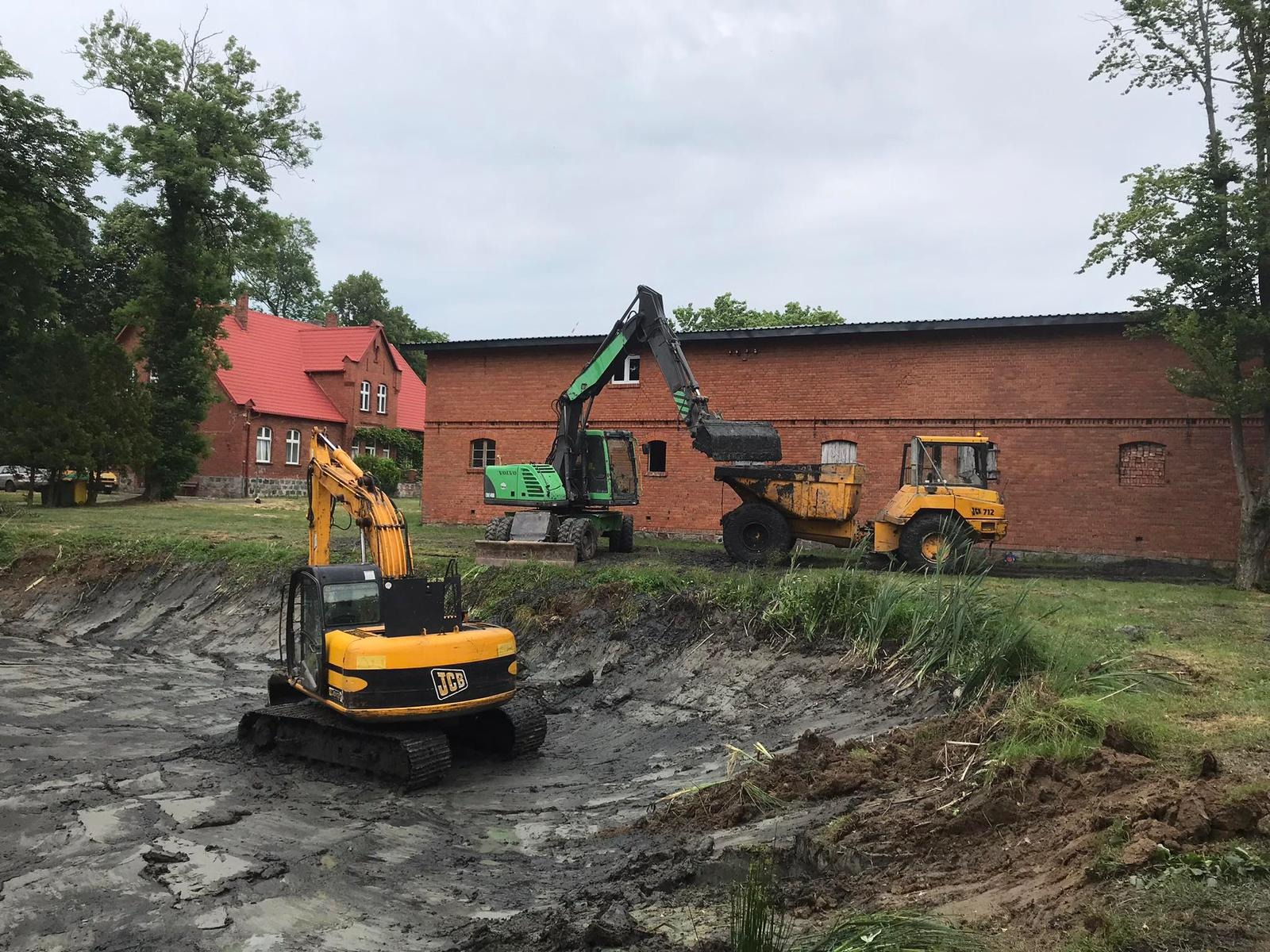 Żółta koparka JCB na pierwszym planie, zielona koparka Volvo ładuje urobek do żółtej wywrotki, w tle budynek z czerwonej cegły i czerwonym dachem, teren po wykopach.