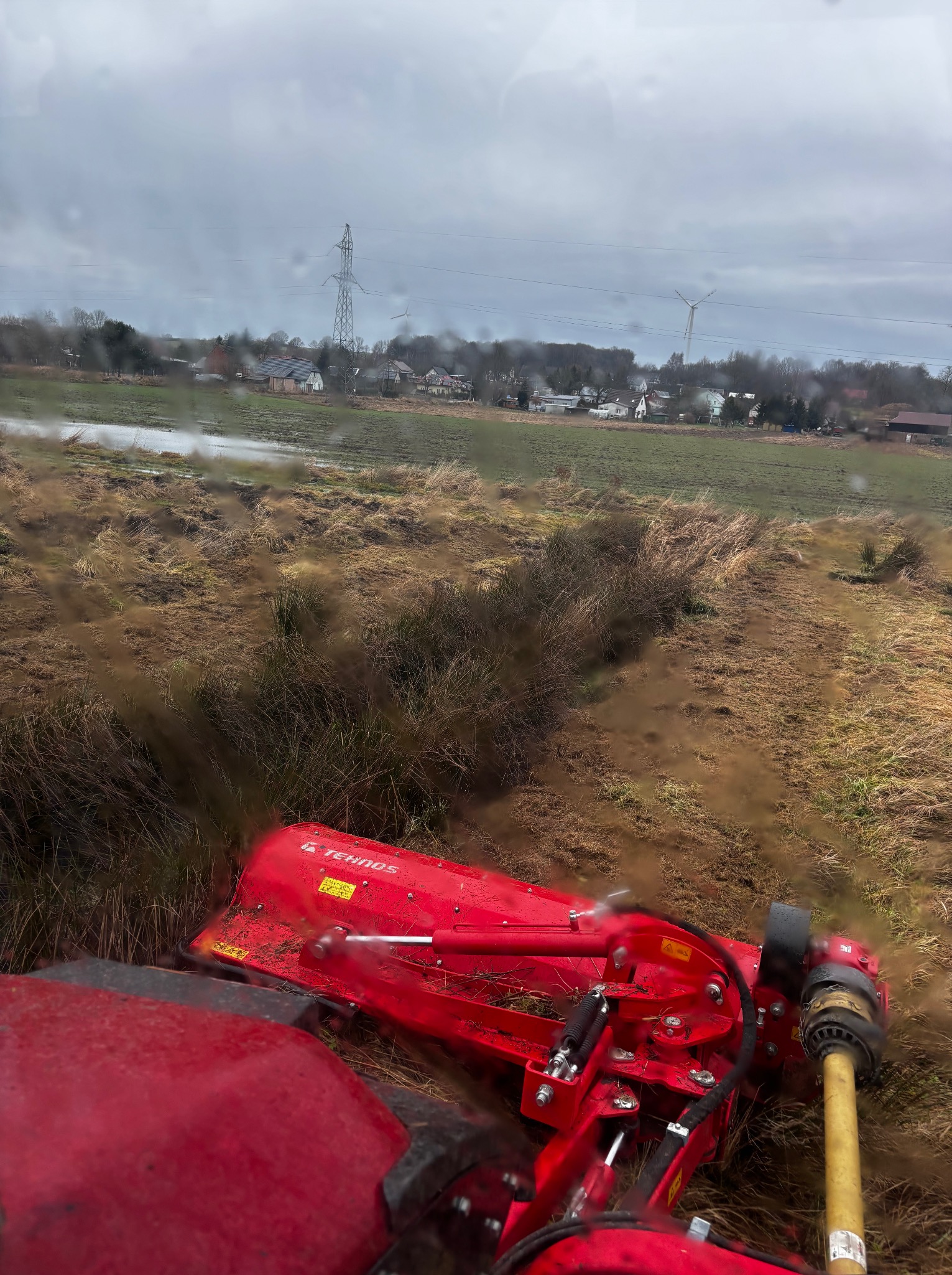 Czerwona kosiarka bijakowa marki Tehnos w trakcie pracy na podmokłej łące, w tle zabudowania wiejskie i słup wysokiego napięcia pod pochmurnym niebem.