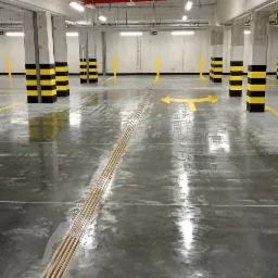 Błyszcząca, mokra posadzka w garażu podziemnym z żółtymi pasami i strzałkami kierunkowymi, czarno-żółte pasy na kolumnach, pomarańczowe rury pod sufitem, równoległe metalowe pręty w podłodze.
