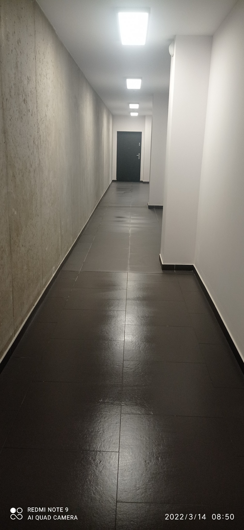 Długi korytarz w budynku mieszkalnym z ciemną podłogą z płytek, oświetlony szeregiem kwadratowych lamp sufitowych, ściany w odcieniach bieli i szarości, widok w perspektywie w kierunku ciemnych...