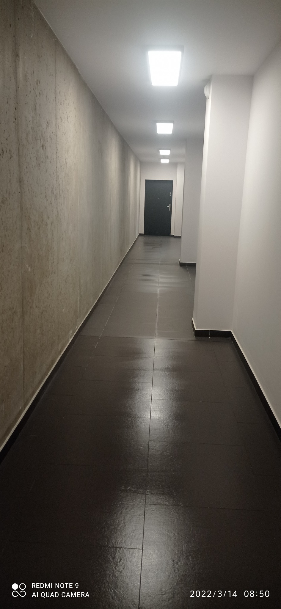 Długi korytarz w budynku z ciemną podłogą z płytek, jasnymi ścianami po prawej i szarą ścianą z fakturą po lewej, zakończony czarnymi drzwiami.
