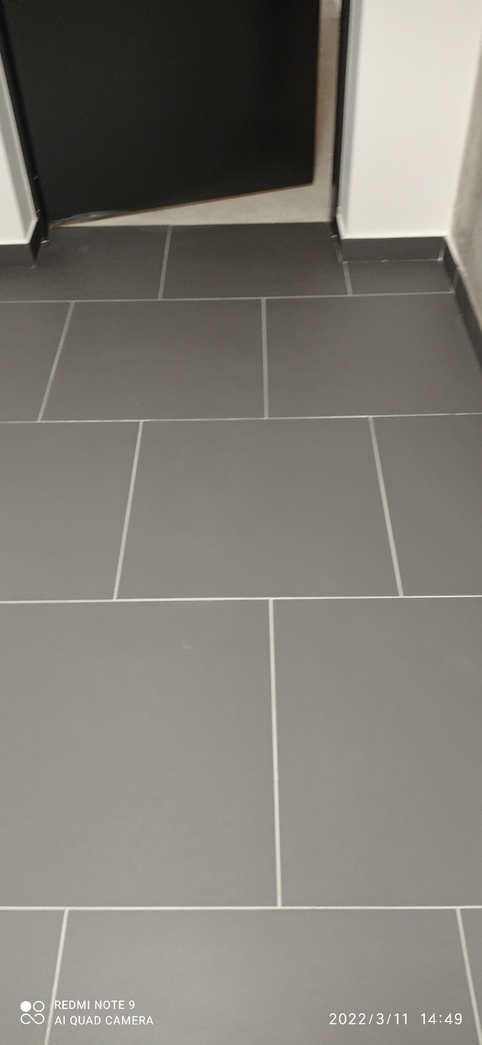 Szare płytki podłogowe ułożone w prostokątnym wzorze, widoczne fragmenty czarnej ościeżnicy drzwi i białej ściany.
