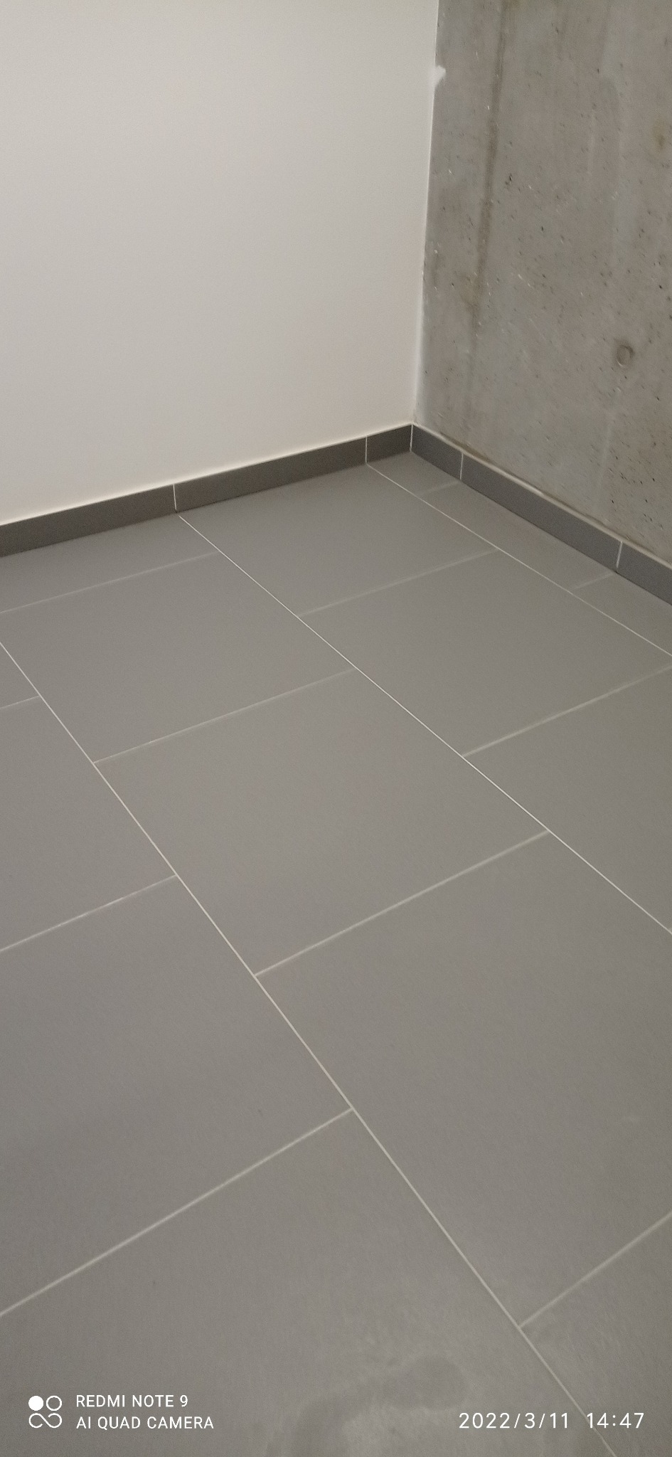 Szare płytki podłogowe w pomieszczeniu z białą i betonową ścianą, widoczne fugi i listwy przypodłogowe.