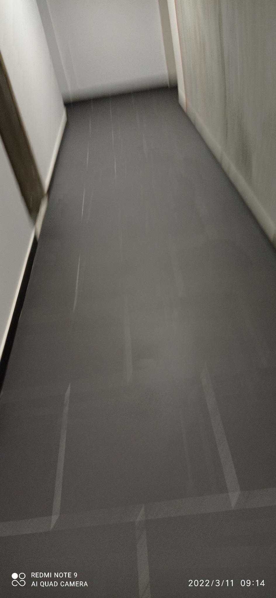 Błyszcząca, ciemna podłoga po umyciu w wąskim korytarzu, widoczne refleksy światła, ściany w odcieniach bieli i szarości.