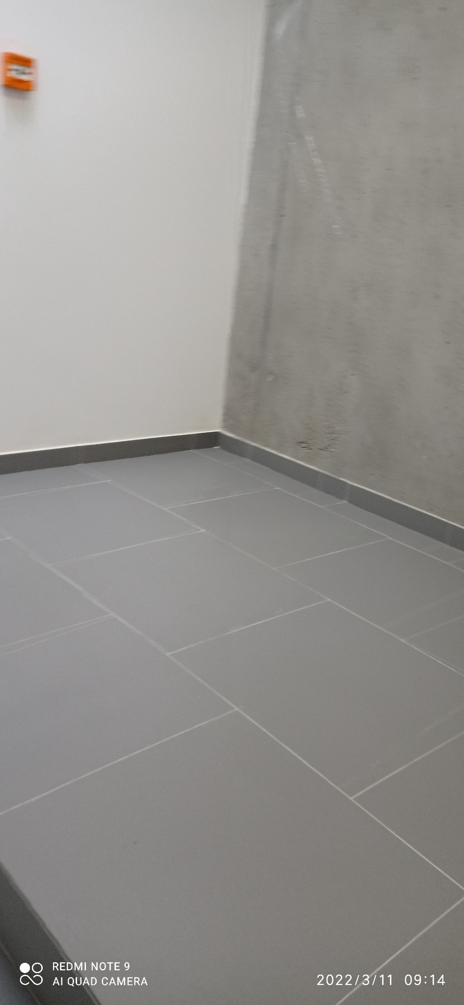 Szare płytki podłogowe w rogu pomieszczenia, ściana biała i surowa szara, widoczny alarm pożarowy na ścianie.