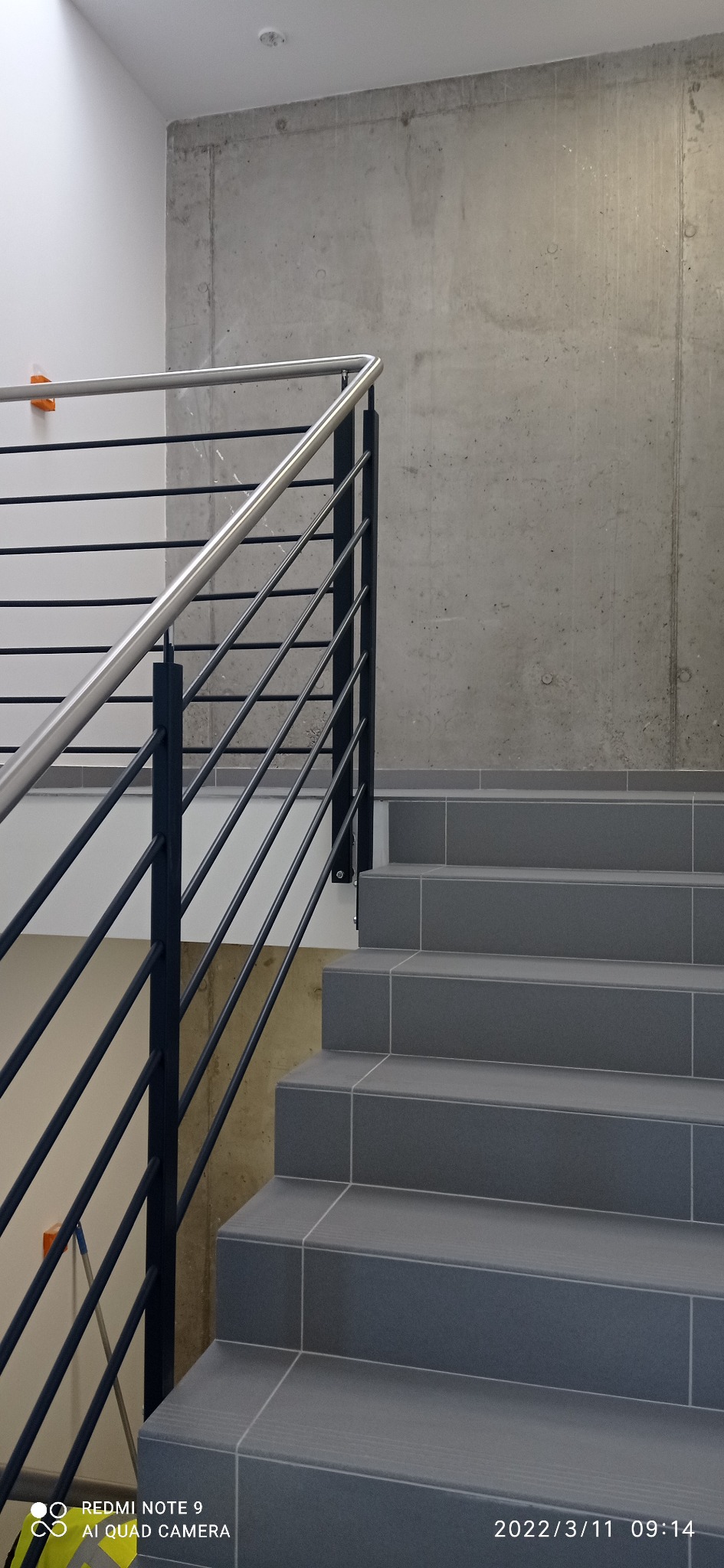 Schody wyłożone szarymi płytkami z metalową balustradą, widoczne ślady po remoncie w postaci zabrudzeń na ścianie i podłodze.