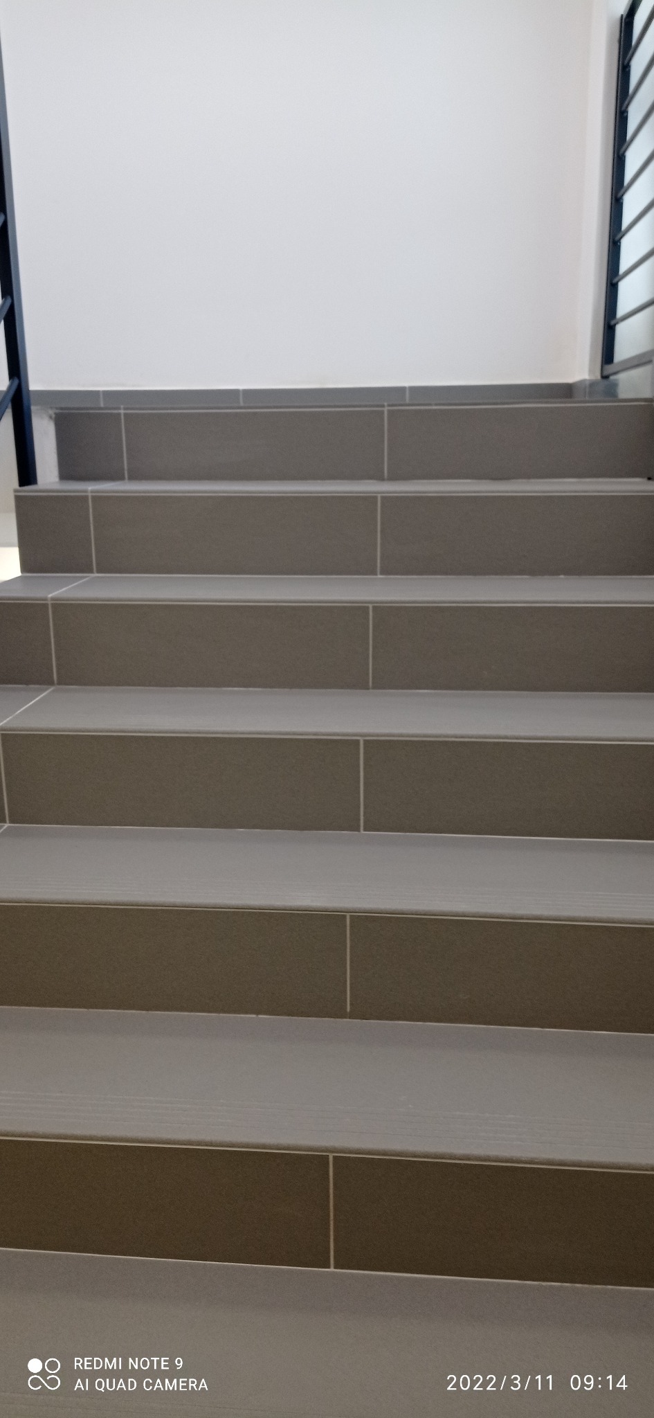 Schody wyłożone szarymi płytkami ceramicznymi z białymi fugami, widok z dołu na górę, fragment balustrady po prawej stronie.