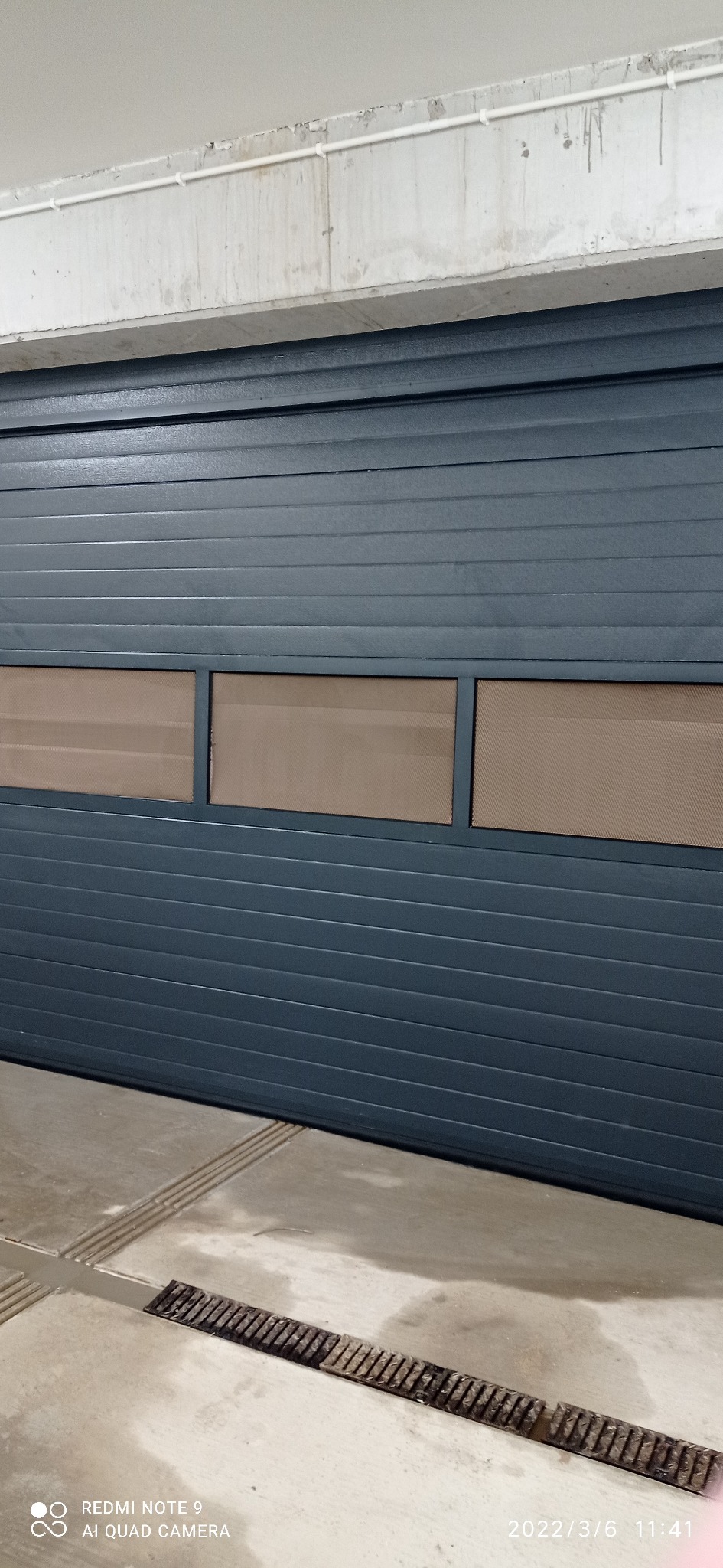 Szara brama garażowa segmentowa z trzema prostokątnymi oknami z siatką, widok na betonową posadzkę z kratką ściekową i fragment betonowego sufitu z rurami.