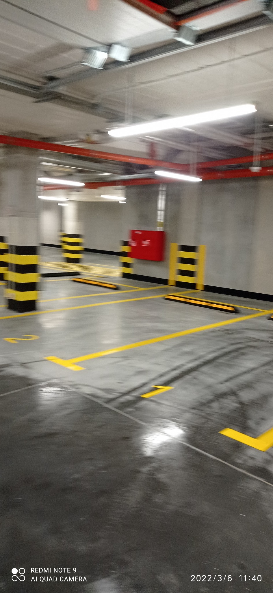 Oznakowane, puste miejsca parkingowe w garażu podziemnym z żółtymi liniami i słupami w czarno-żółte pasy, widoczna instalacja przeciwpożarowa.