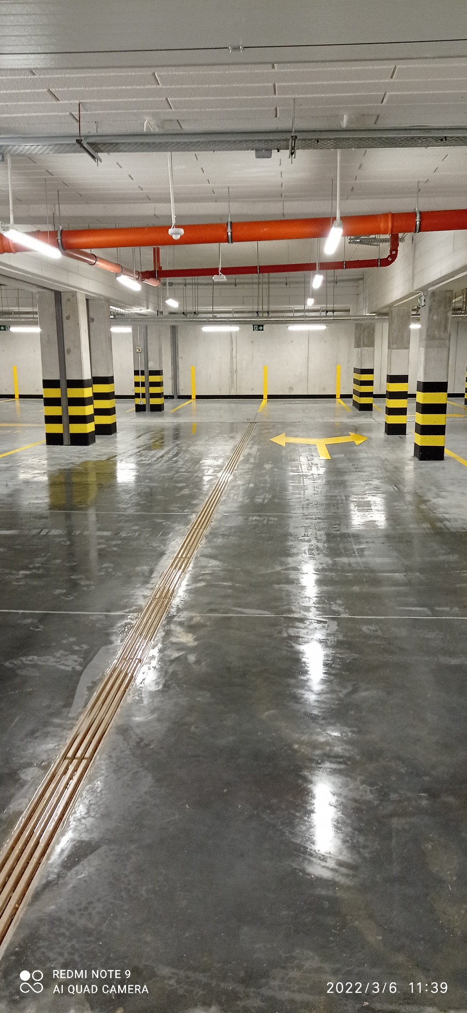 Błyszcząca, mokra posadzka w garażu podziemnym z żółtymi pasami i strzałkami kierunkowymi, czarno-żółte pasy na kolumnach, pomarańczowe rury pod sufitem, równoległe metalowe pręty w podłodze.