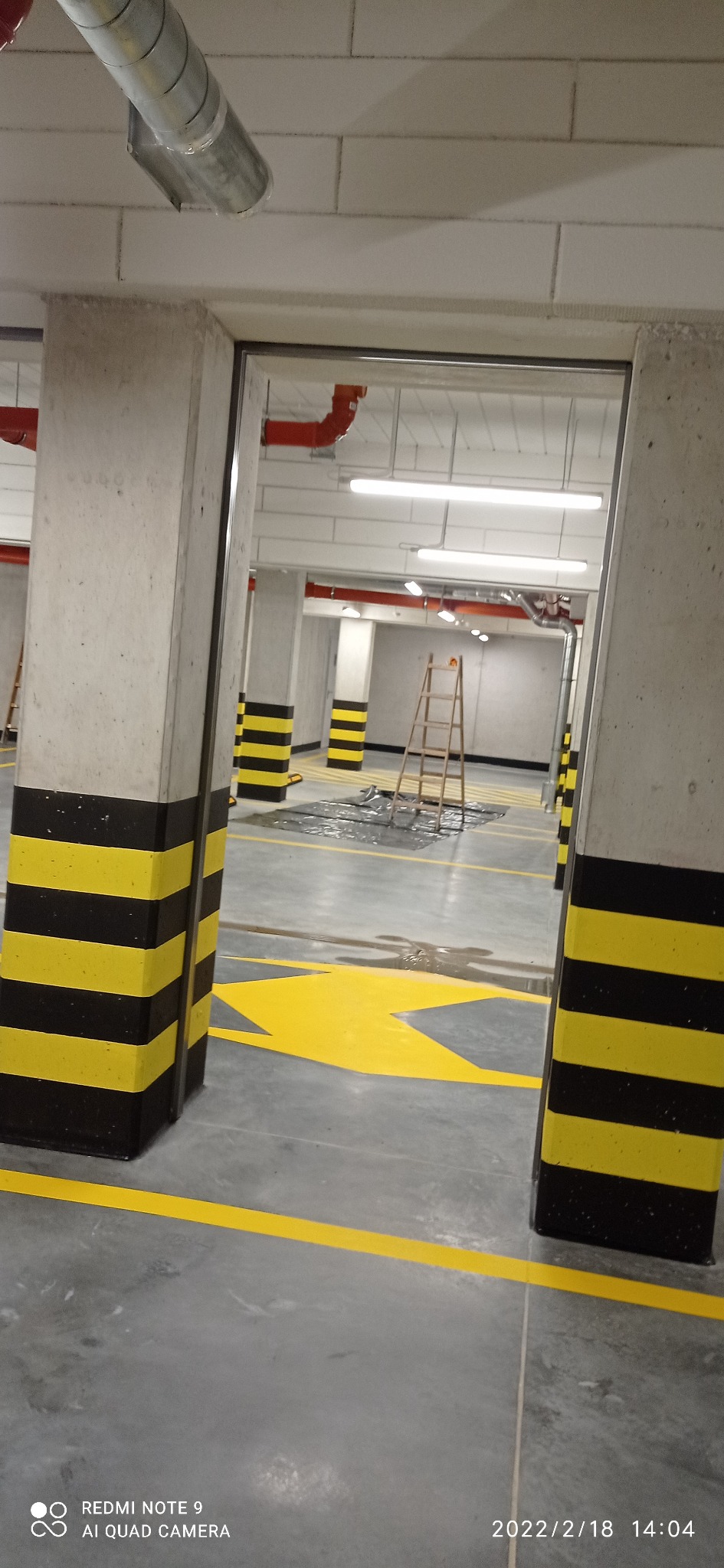 Oznakowanie poziome w garażu podziemnym: świeżo pomalowane żółte strzałki i pasy na szarej betonowej posadzce, kolumny z czarno-żółtymi pasami ostrzegawczymi, drabina w tle, widoczne ślady mokrej...