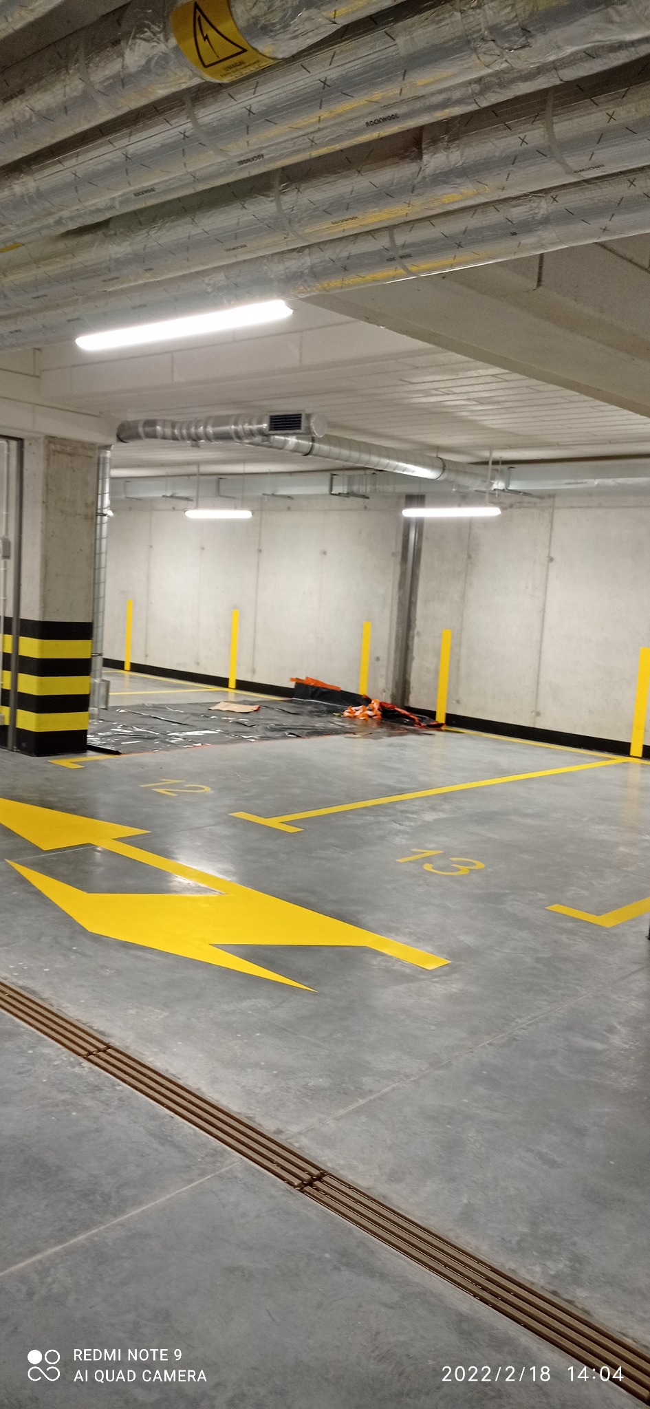 Oznakowane żółtą farbą stanowiska parkingowe w podziemnym garażu, widoczne rury wentylacyjne pod sufitem i słup z czarno-żółtymi pasami.