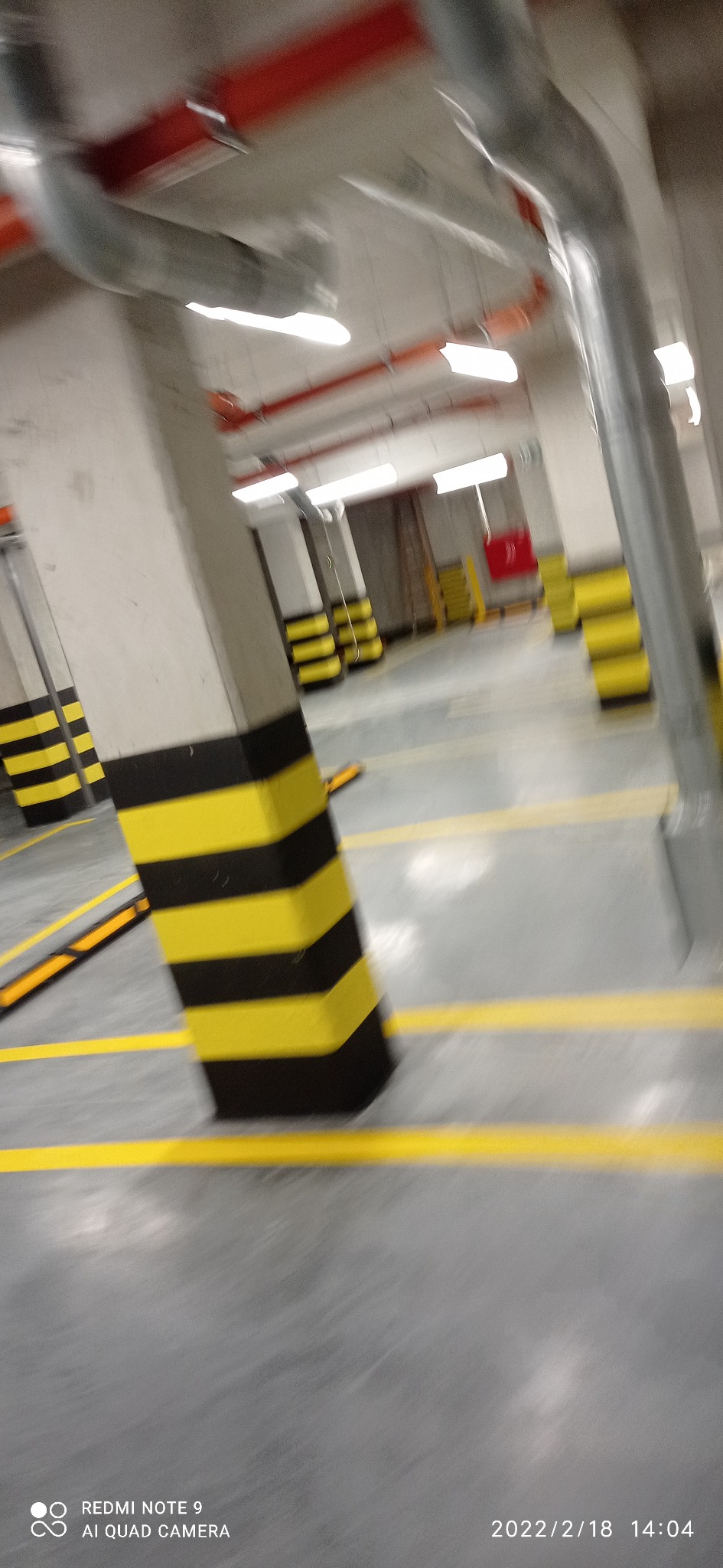 Wnętrze parkingu podziemnego z betonowymi kolumnami w czarno-żółte pasy, żółtymi liniami na podłodze i widocznymi instalacjami pod sufitem.
