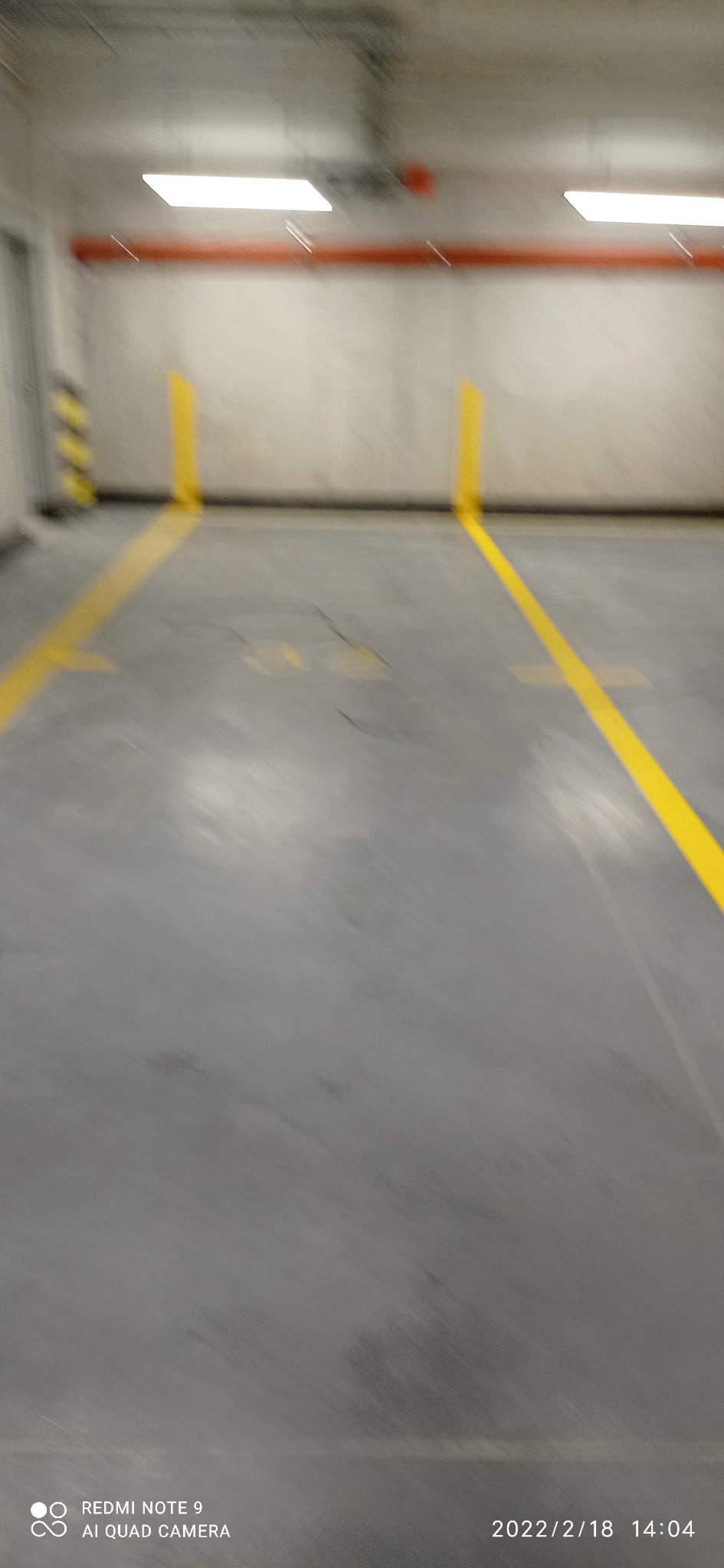 Szare podłoże z żółtymi liniami wyznaczającymi ciągi komunikacyjne w hali lub magazynie, widoczny fragment ściany z oznaczeniami ostrzegawczymi.