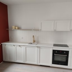 Daniel Wiśniewski - Nowoczesna kuchnia z białymi szafkami, złotą armaturą i ceglastą ścianą w tle, prezentująca minimalistyczny design po remoncie.