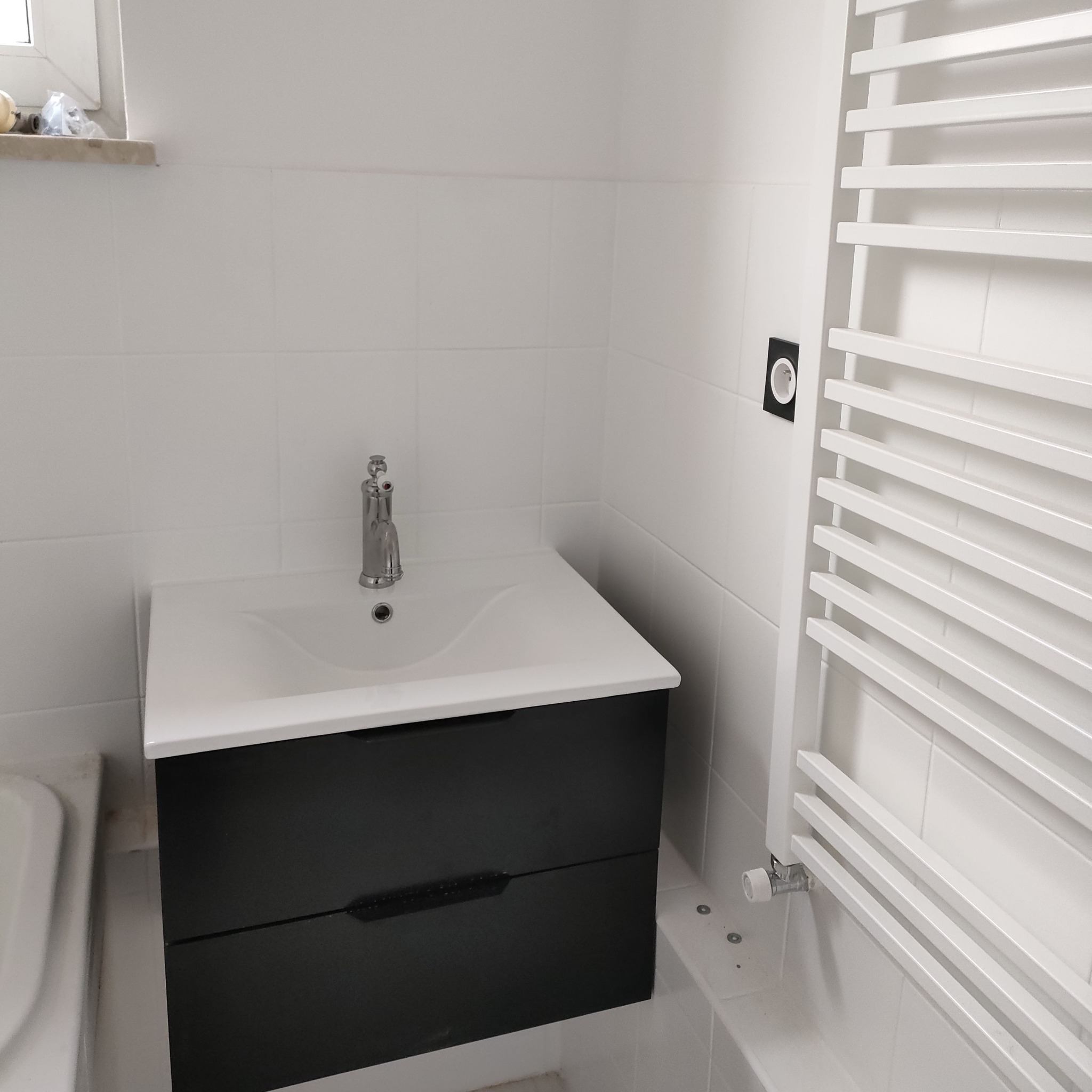 Biała łazienka z czarną szafką pod umywalką, chromowaną baterią i białym grzejnikiem drabinkowym na tle białych płytek.