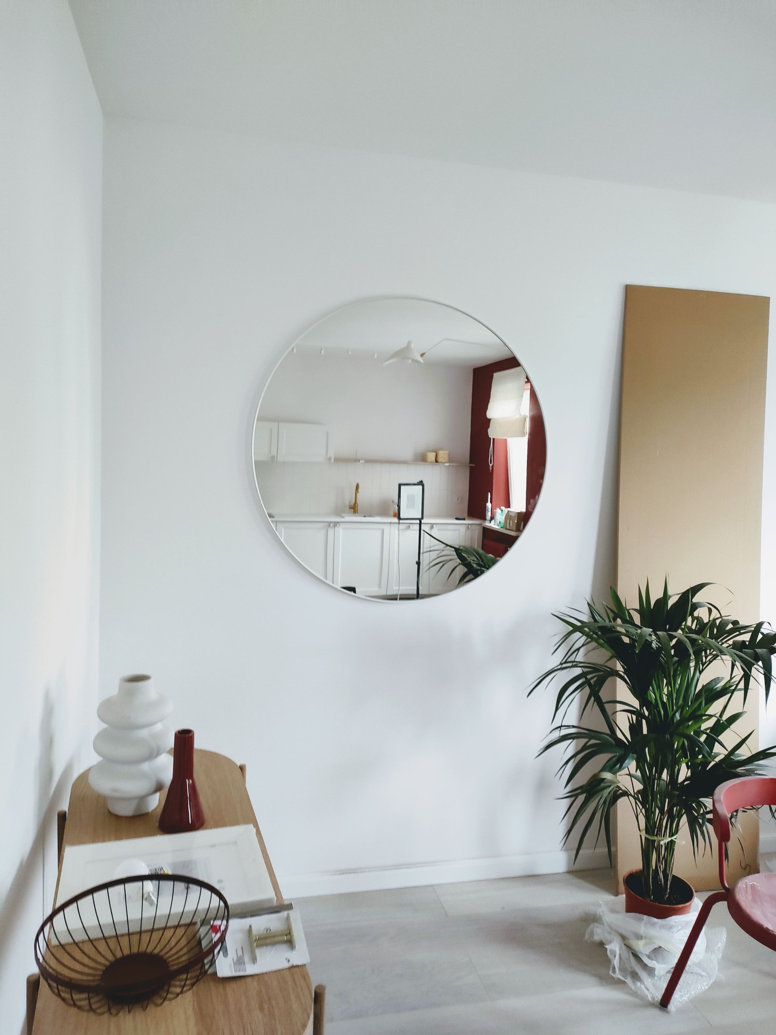 Jasne wnętrze z okrągłym lustrem odbijającym kuchnię z białymi szafkami i czerwoną ścianą, roślina doniczkowa obok panelu, stolik z dekoracjami.