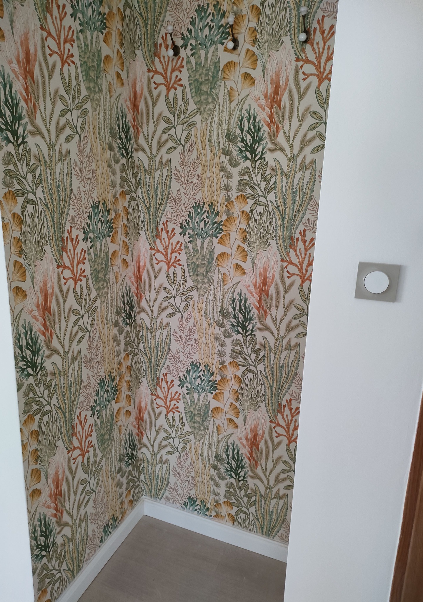 Wnętrze z tapetą w motywy roślinne, imitujące podwodną florę w odcieniach zieleni, brązu i pomarańczy; widoczny fragment ściany z włącznikiem światła oraz narożnik pokoju z listwą przypodłogową.