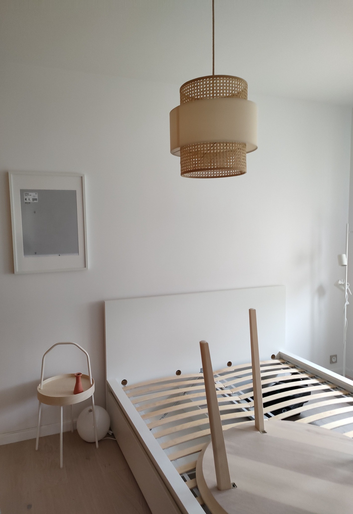 Wnętrze pokoju z białymi ścianami, zdemontowane łóżko z drewnianym stelażem, stolik nocny z dekoracyjnym wazonem i wisząca lampa z elementami rattanowymi.