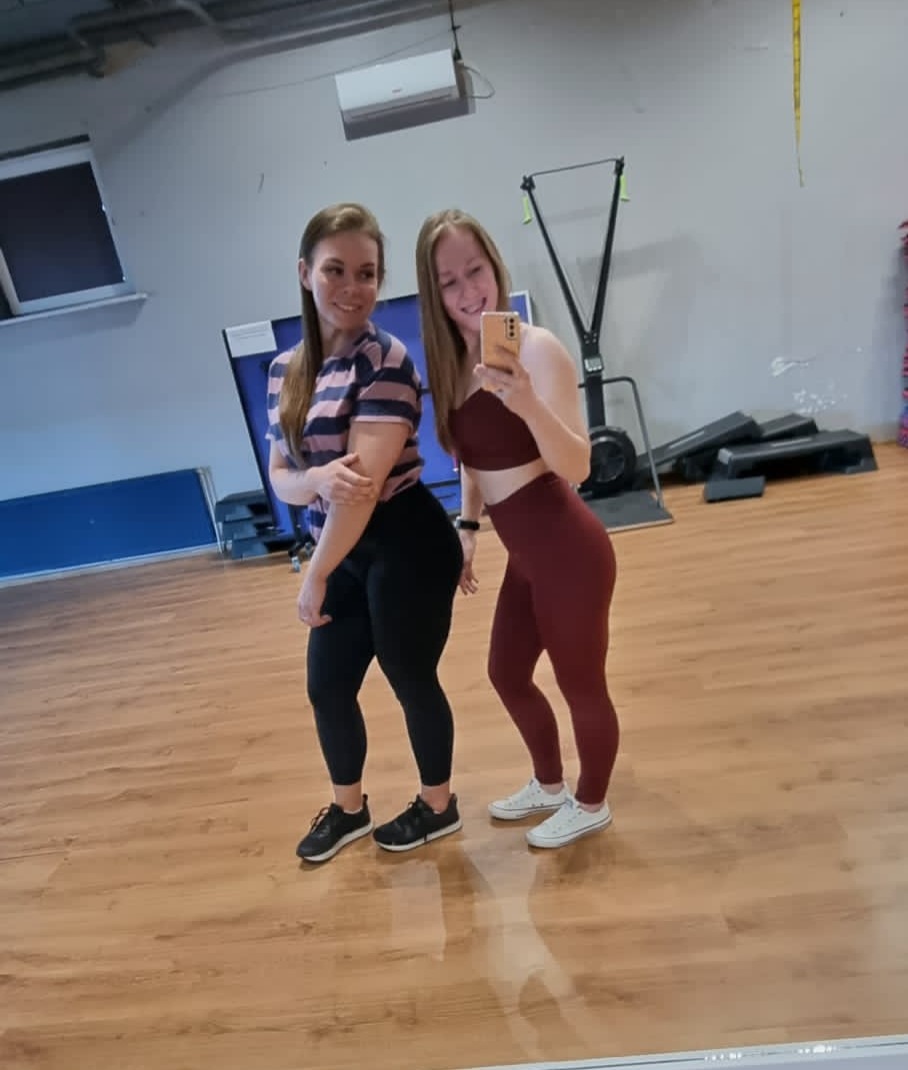 Dwie uśmiechnięte kobiety w strojach sportowych pozują do selfie w siłowni z drewnianą podłogą i sprzętem do ćwiczeń w tle.