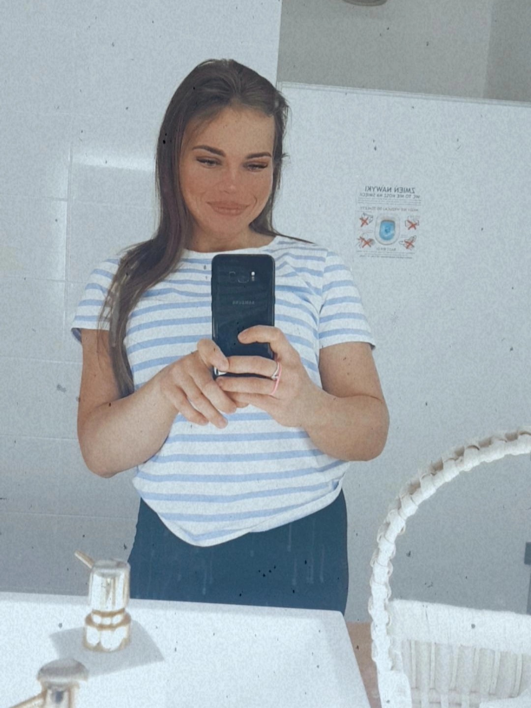 Kobieta w paski robi selfie w łazience, widoczny fragment kosza wiklinowego i naklejka informacyjna na ścianie.